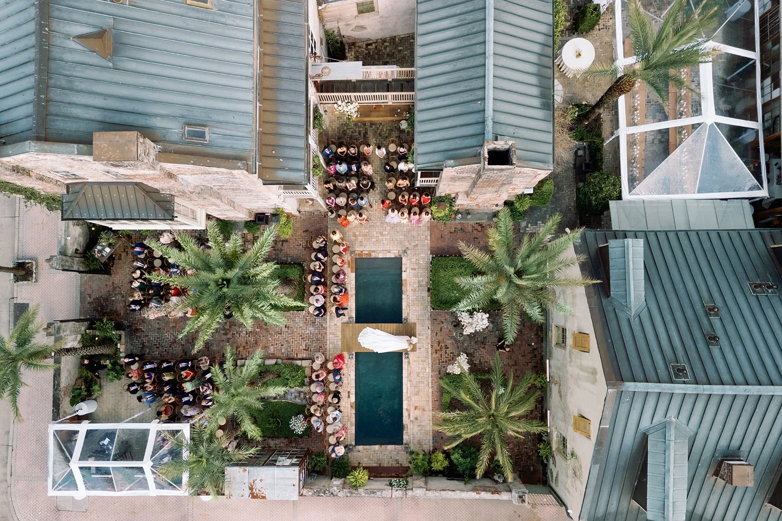 An aerial view of a courtyard wedding ceremony, with a bride and groom at an altar, guests seated on either side, tall palm trees, and surrounding buildings with rooftops and glass enclosures.
