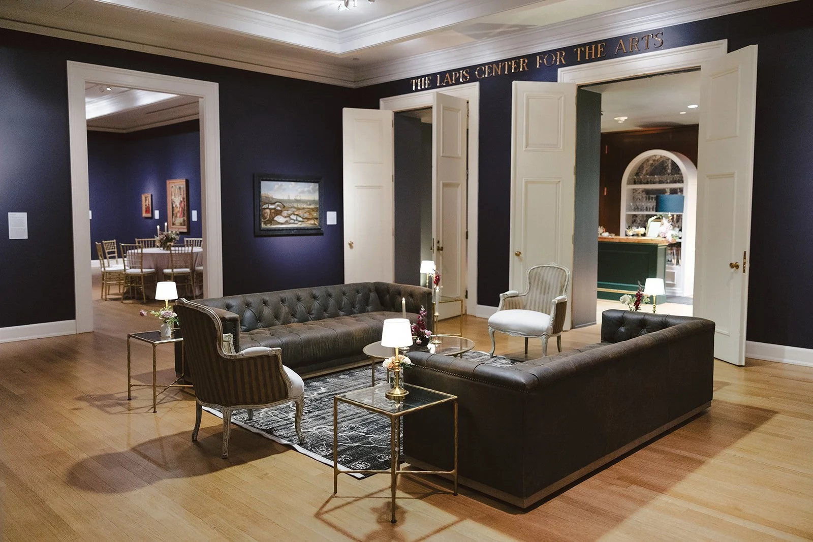 A living room with dark blue walls, hardwood floors, and white trim. It contains a gray tufted sofa, white armchair, and small side tables with lamps. There is a black and white rug and artwork on the walls. Double doors open to another room, and a s