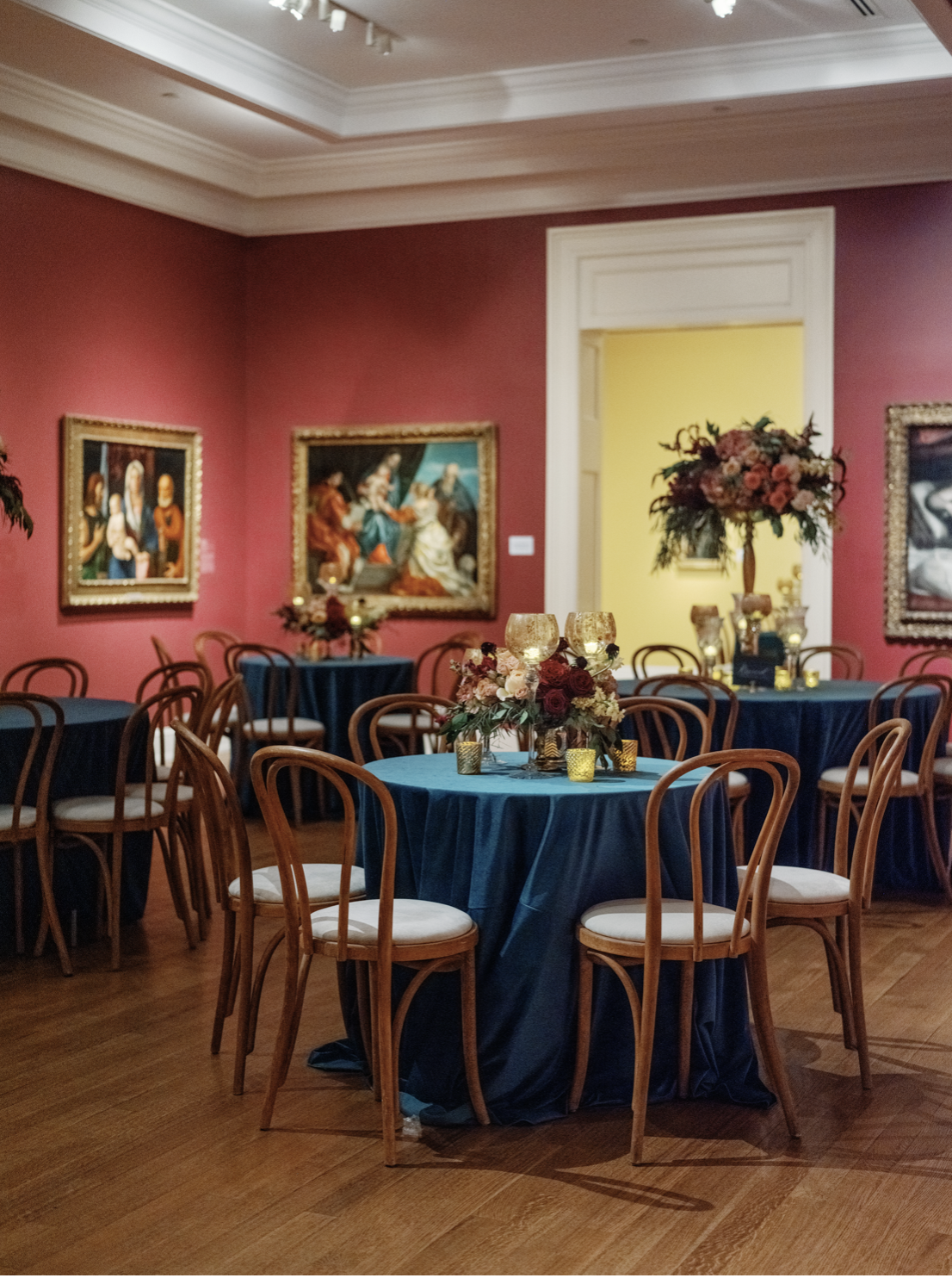 Elegant dining area with round tables draped in dark blue tablecloths, decorated with floral centerpieces and candle holders, in a room with pink walls adorned with classical paintings.