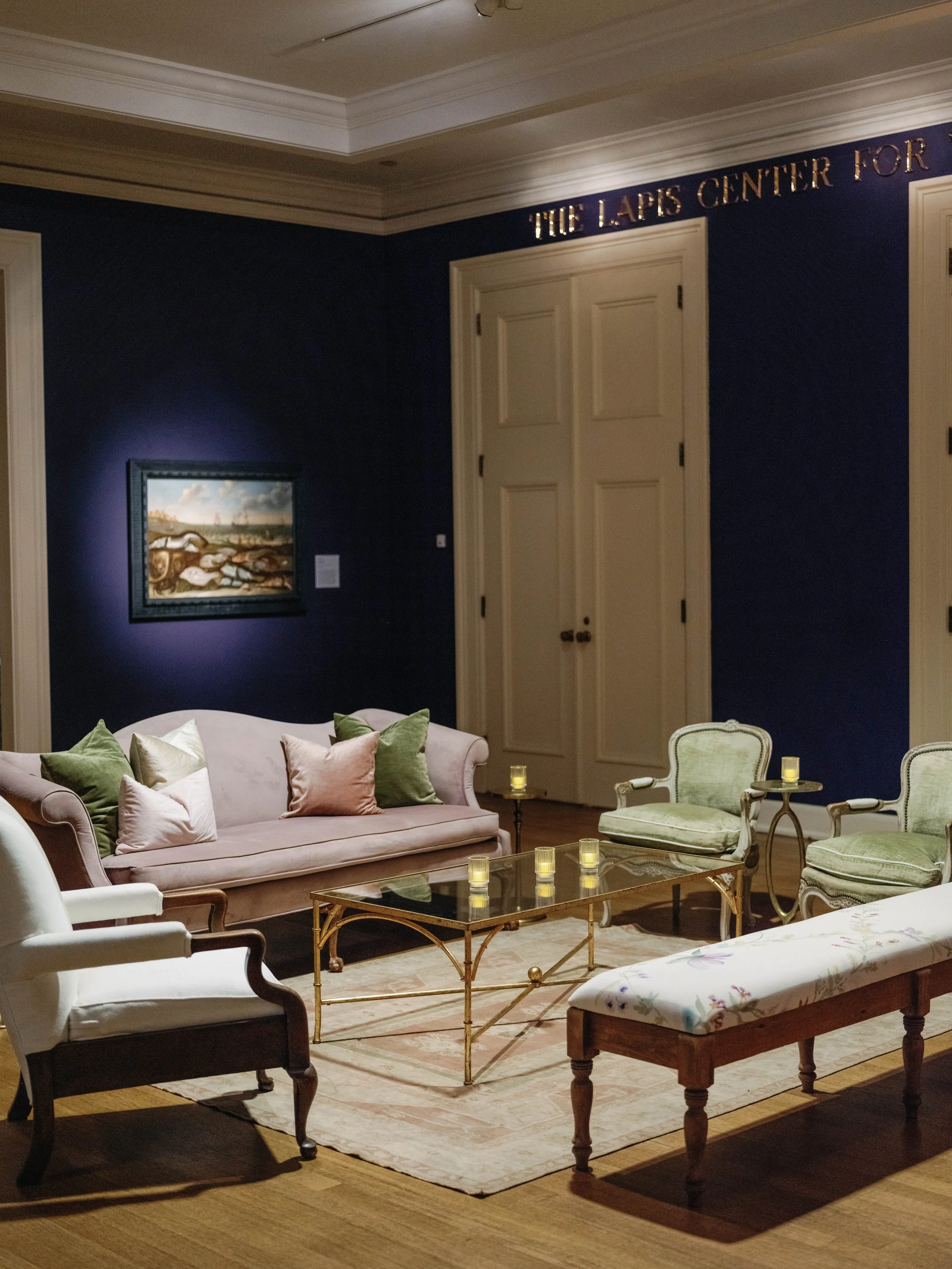 A living room with a pink upholstered sofa with green and pink pillows, a glass-topped gold coffee table with candles, white and green chairs, a framed painting of a landscape, and a cream-colored bench with a floral cushion, against a navy blue wall