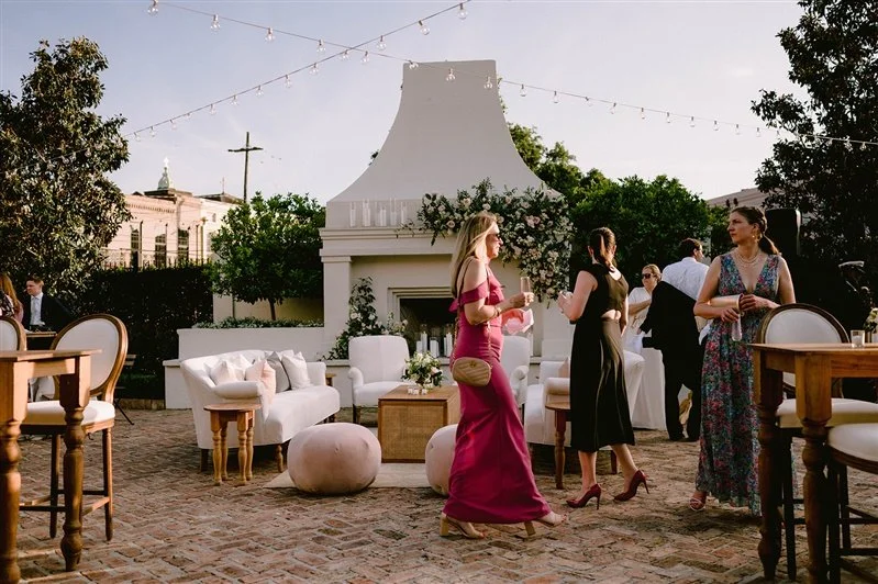 People socializing at an outdoor party with white furniture, string lights, and a large white fireplace with floral decorations.