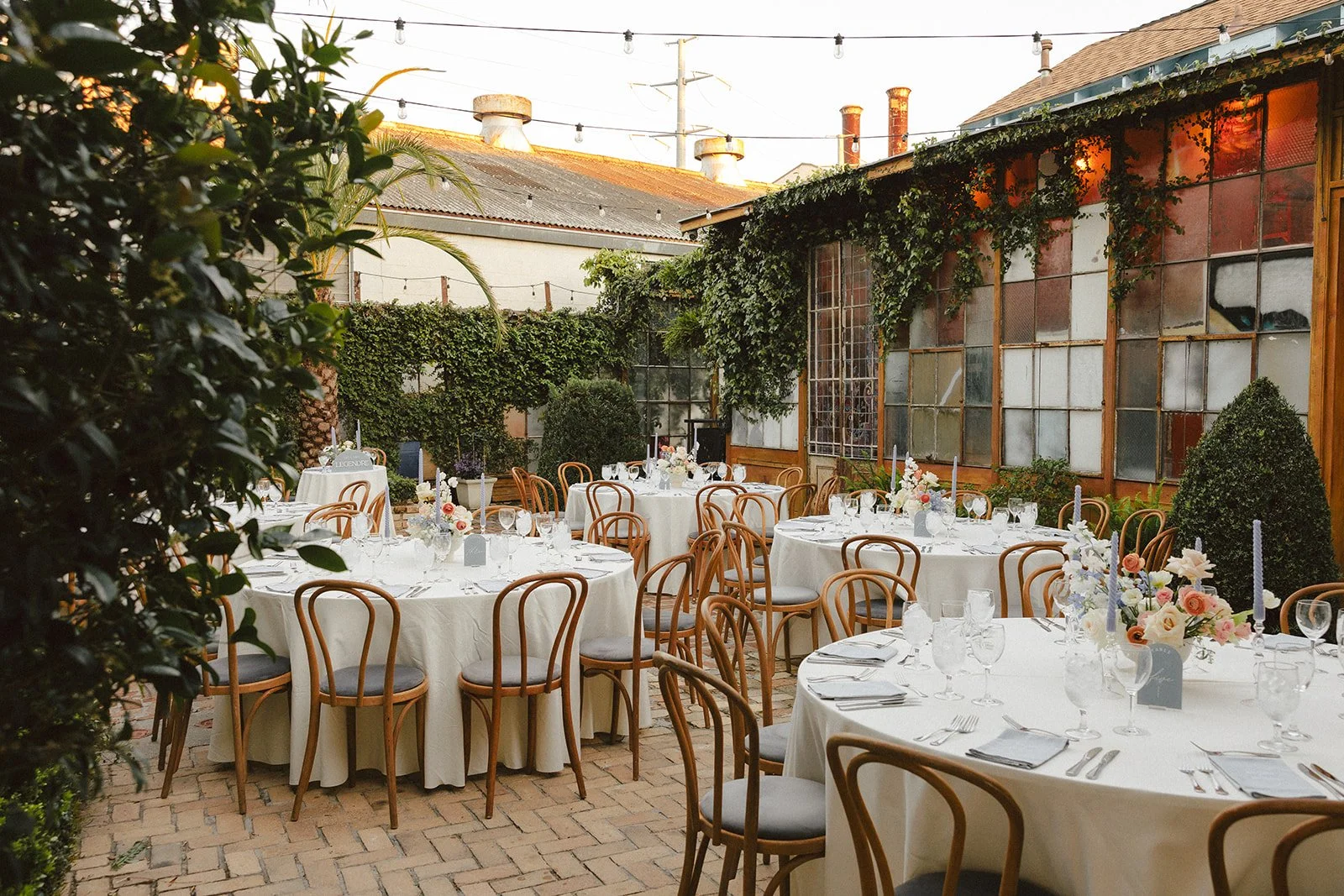 Outdoor event space with round tables draped in white tablecloths, decorated with floral centerpieces and candles, set up for a formal dinner under string lights, surrounded by greenery and a rustic building with large windows.