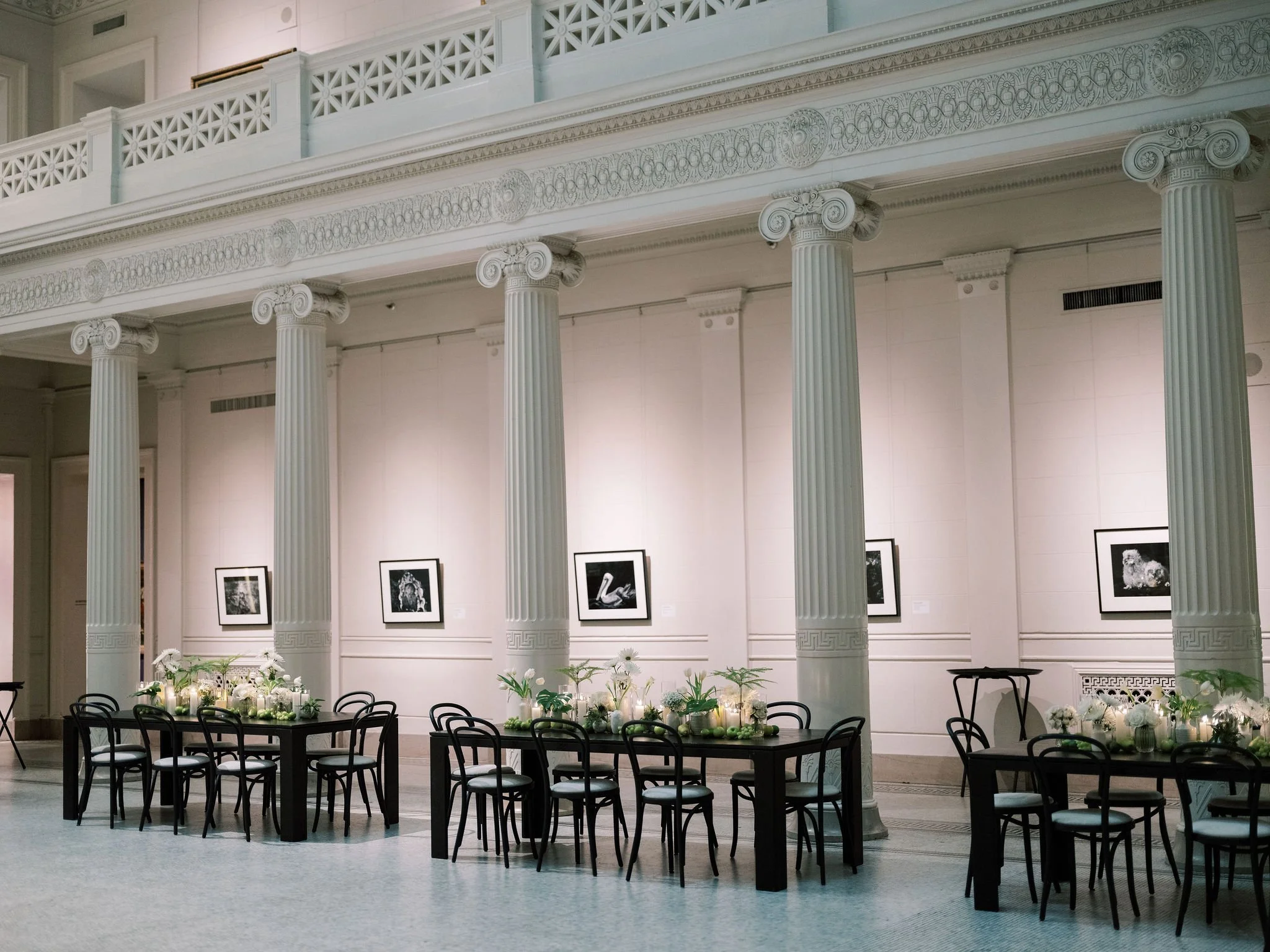 Interior of a grand space with tall, ornate Ionic columns supporting a decorative entablature with intricate designs. Walls are adorned with framed black-and-white artwork. A table in the foreground is decorated with white flowers and green candles, 