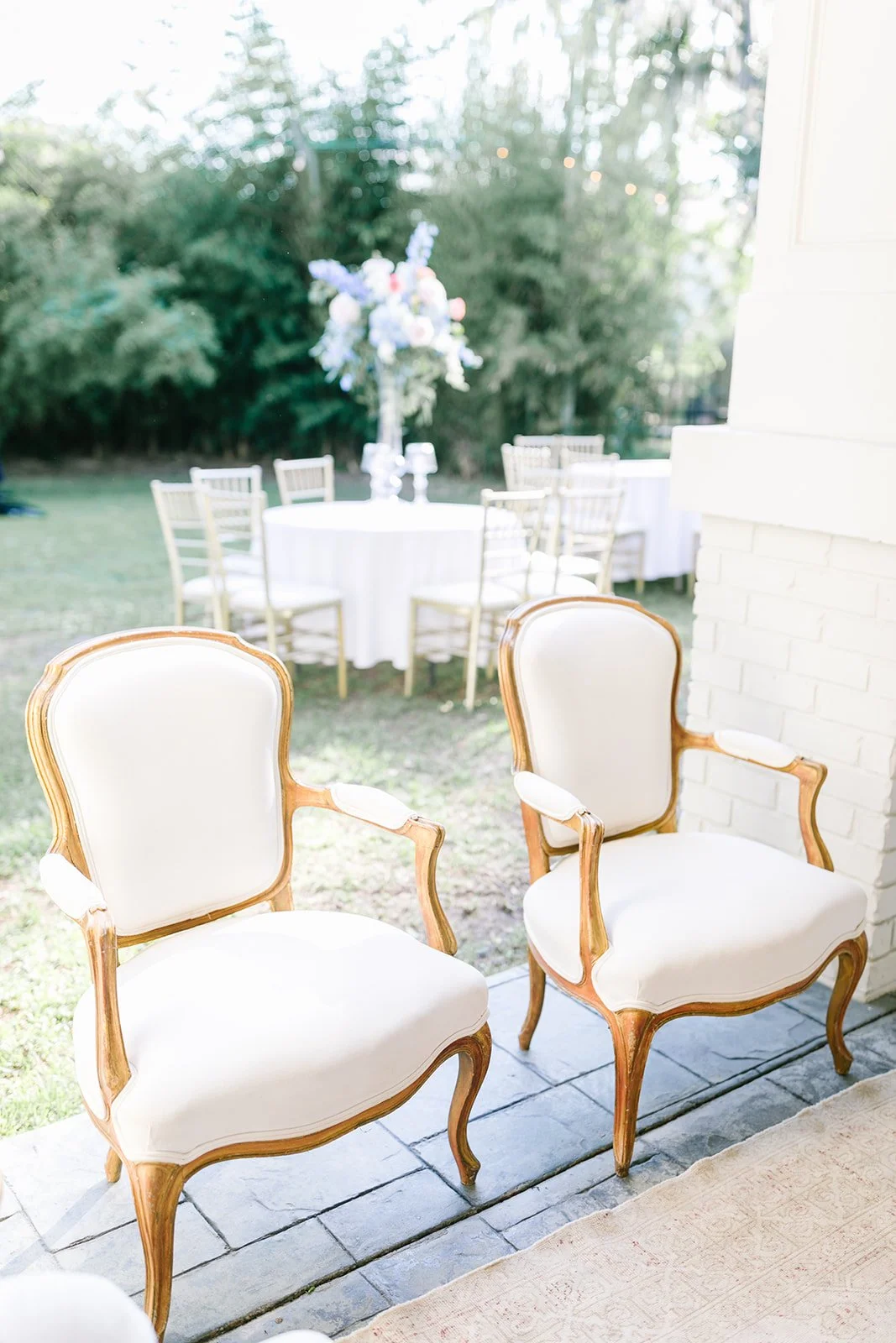 Two vintage-style white armchairs with wooden frames are placed on a stone patio near a white brick wall, overlooking an outdoor garden with round tables and gold chairs, set for an event with a floral centerpiece in the background.