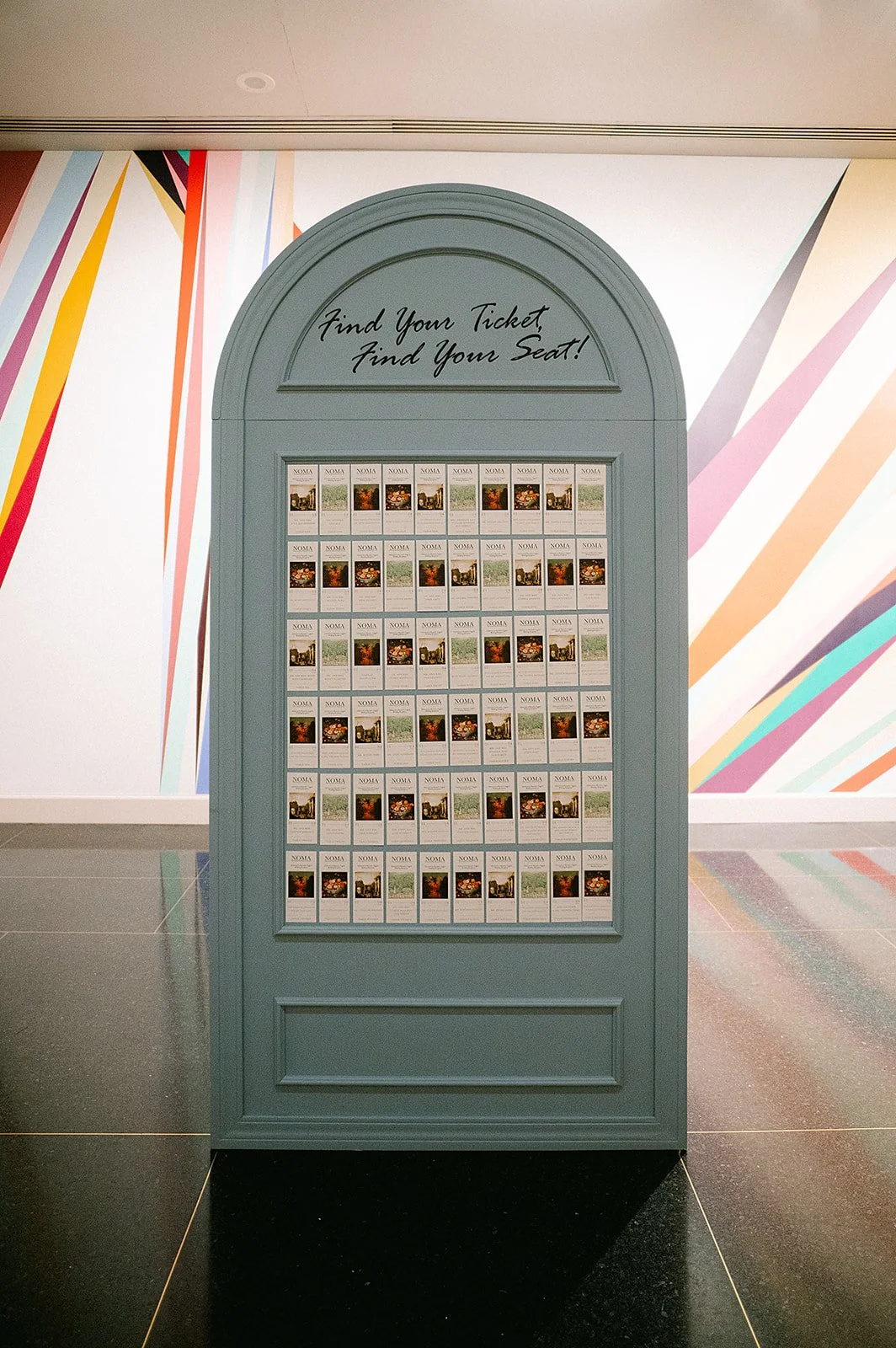 A blue ticket display board with multiple tickets inside, and a handwritten sign at the top that says "Find Your Ticket, Find Your Seat!" The background features colorful diagonal lines painted on a white wall.