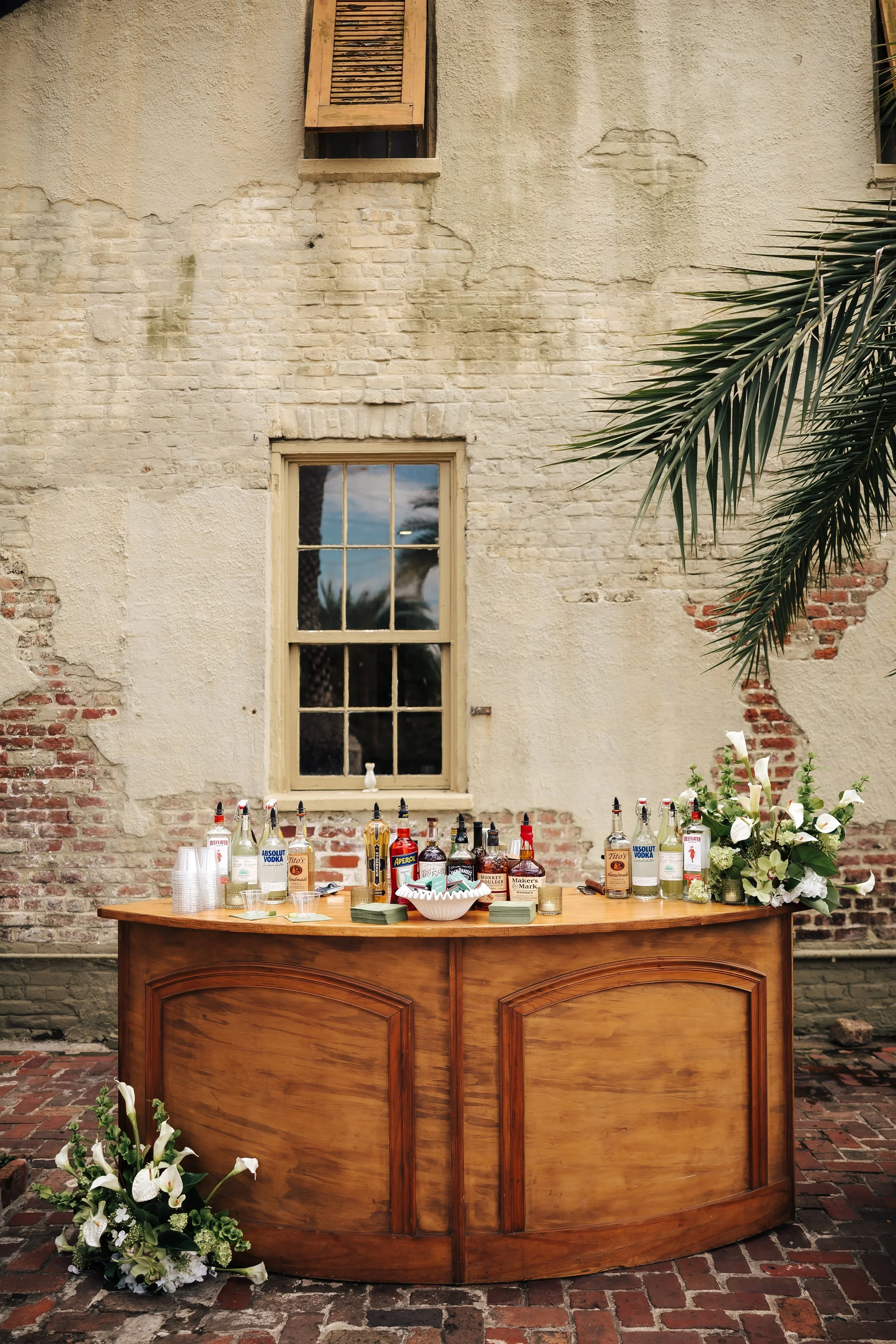 A wooden bar table set up outdoors against an old brick and stucco wall with a window. The table has various liquor bottles, plastic cups, napkins, and floral arrangements with white flowers and greenery.