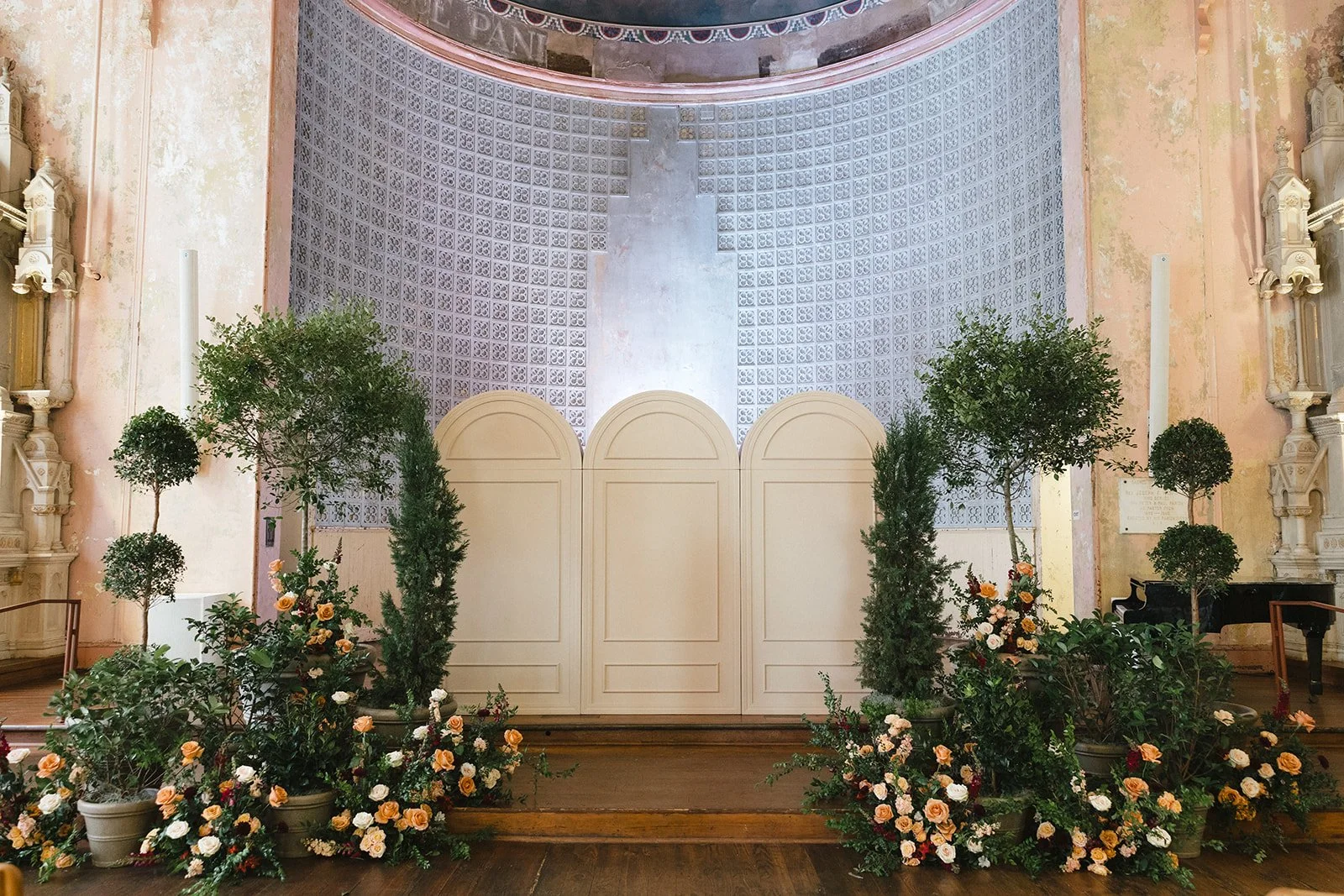 Indoor wedding or event altar decorated with potted plants and flowers, with a white wooden panel and ornate wall features in the background.