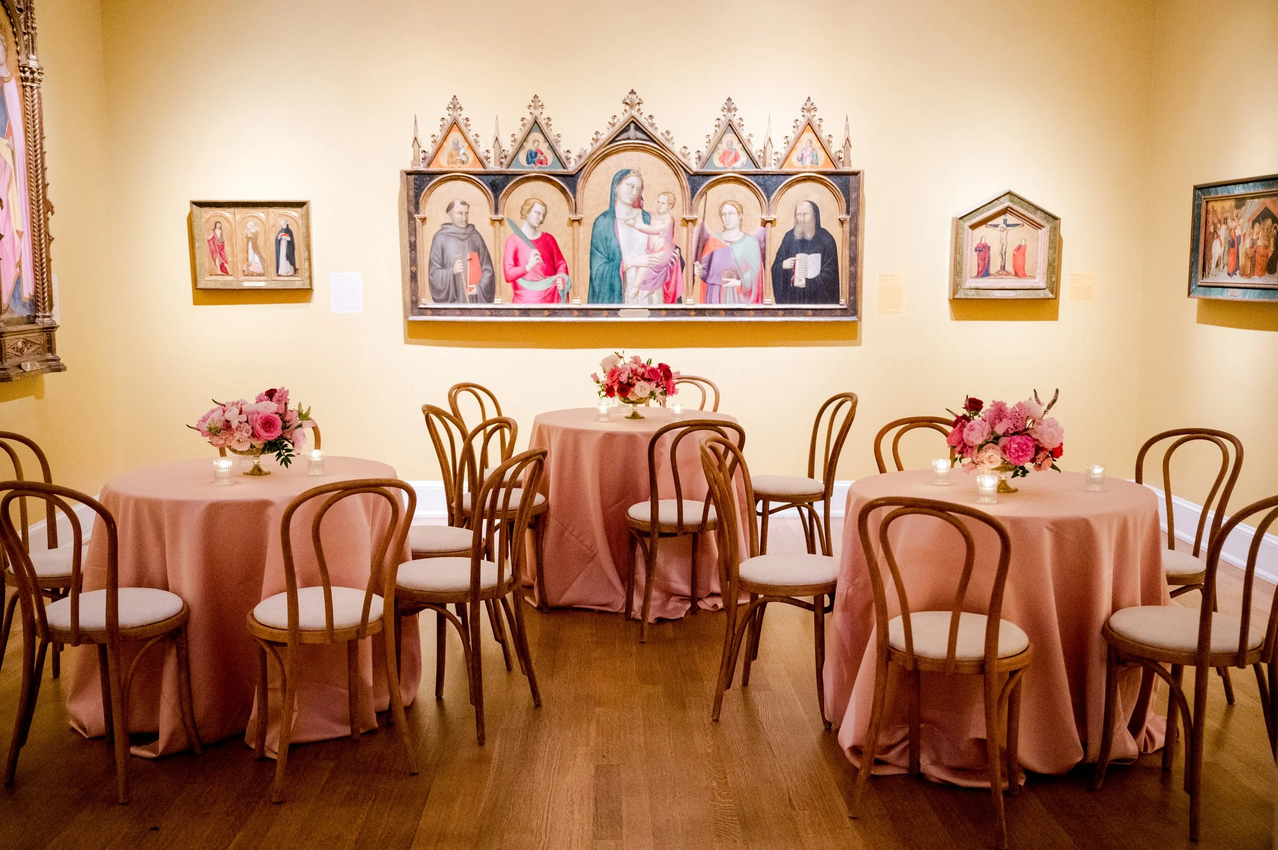 A banquet room with round tables covered in pink tablecloths, each decorated with a bouquet of pink and white flowers; there are wooden chairs around the tables, and the wall behind features religious artwork including a large triptych of saints and 