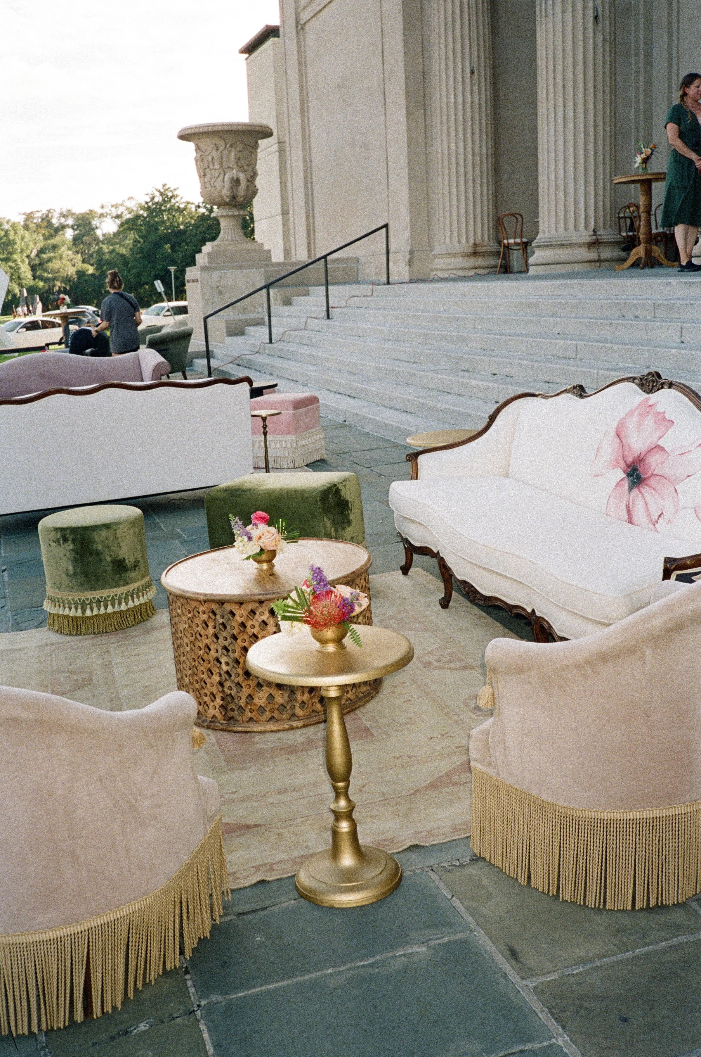 An outdoor event setup featuring vintage-style furniture and decor. There are sofas with fringed skirts, small round tables with floral arrangements, and a set of steps leading up to a building with large columns. Several people are visible, one stan