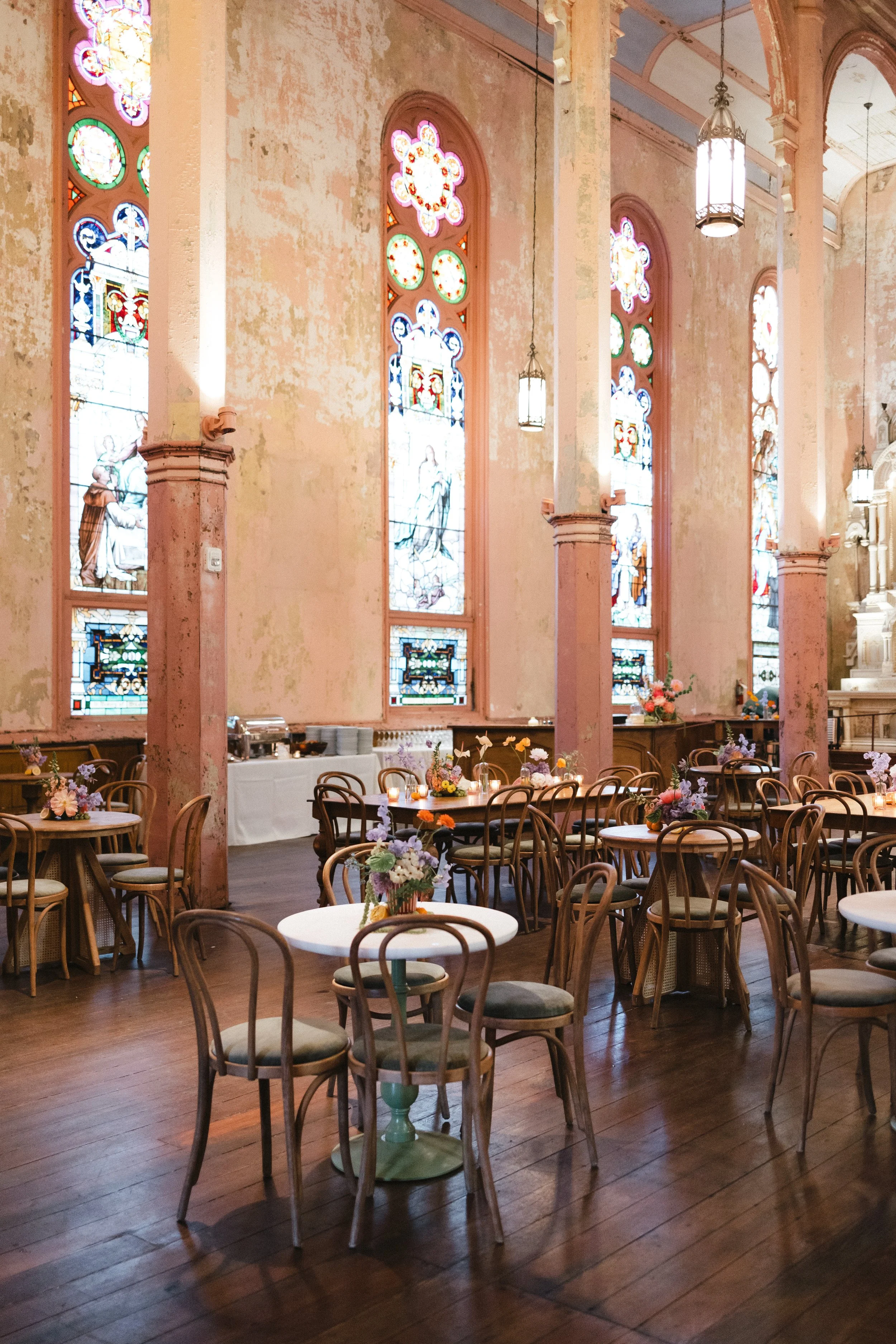 Interior of an old church with stained glass windows, wooden tables and chairs with floral arrangements for a gathering or celebration.
