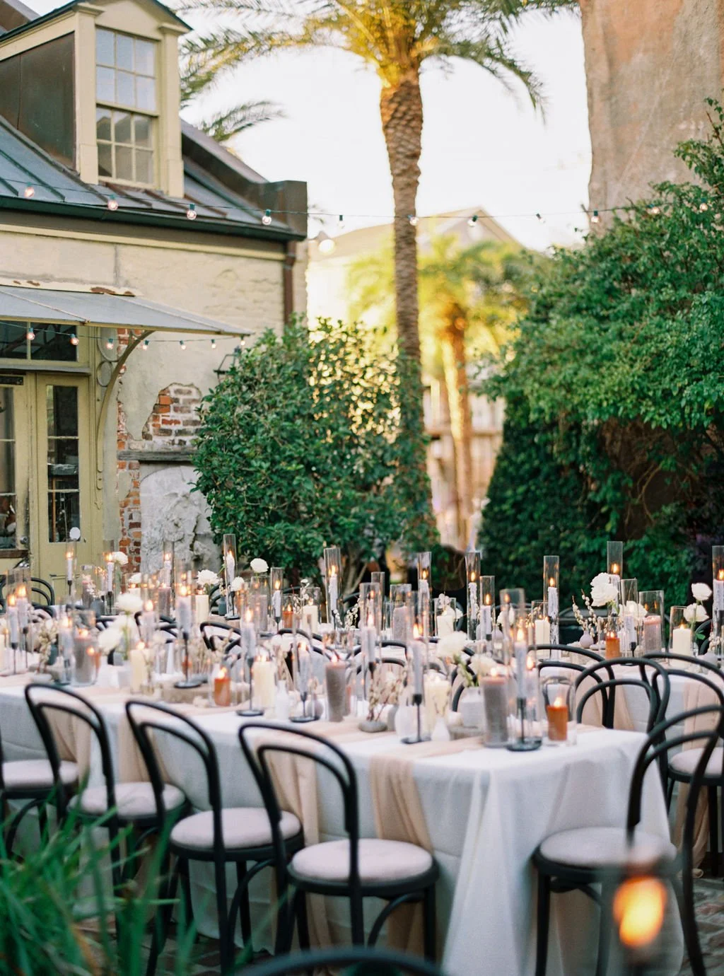 Outdoor dining setup with tables, white tablecloths, and candles, surrounded by lush greenery and string lights, in a garden setting during sunset.