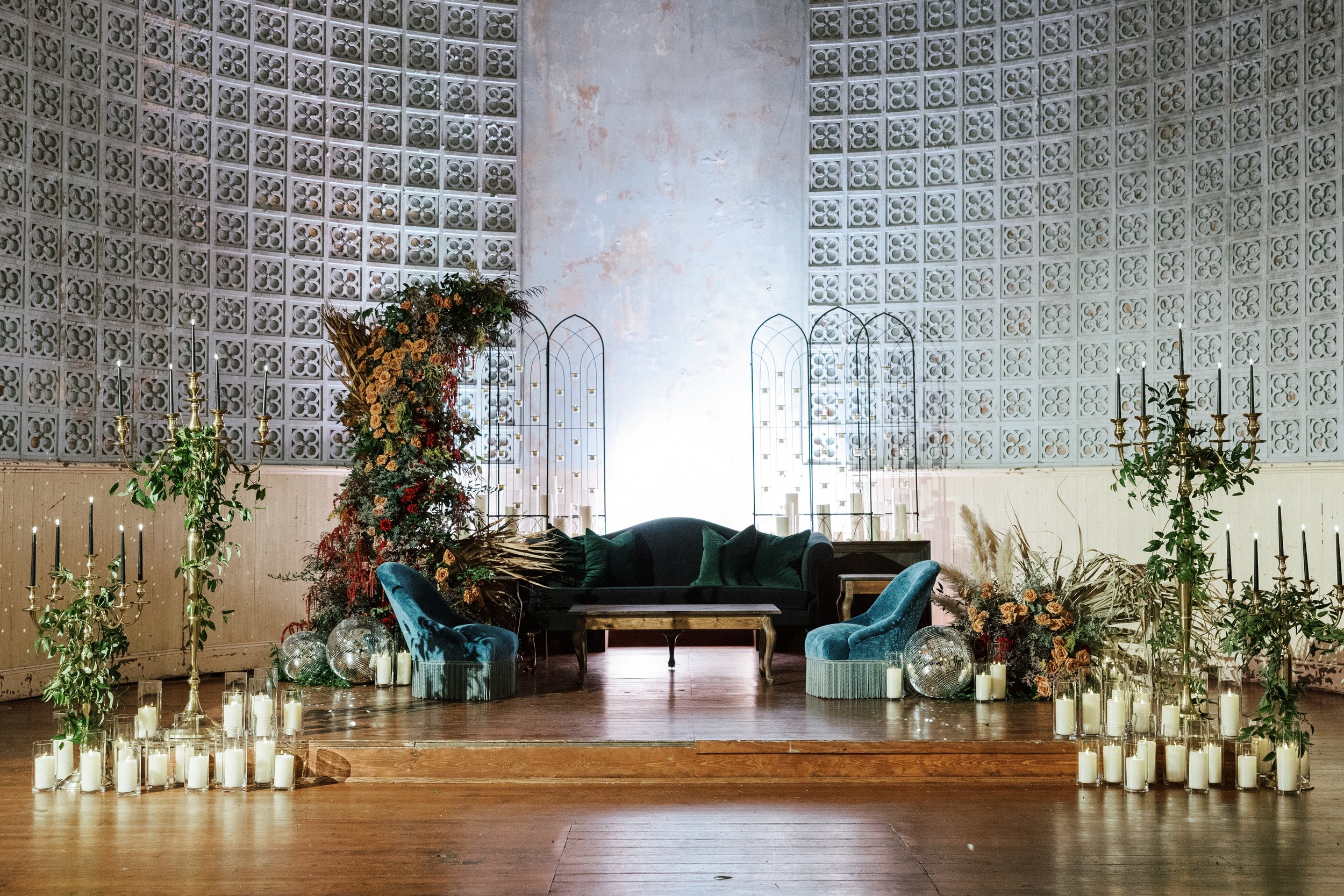 Interior of a decorated event space with a floral arrangement, green plants, candle holders, and candles on the floor, against a textured wall with lattice panels.