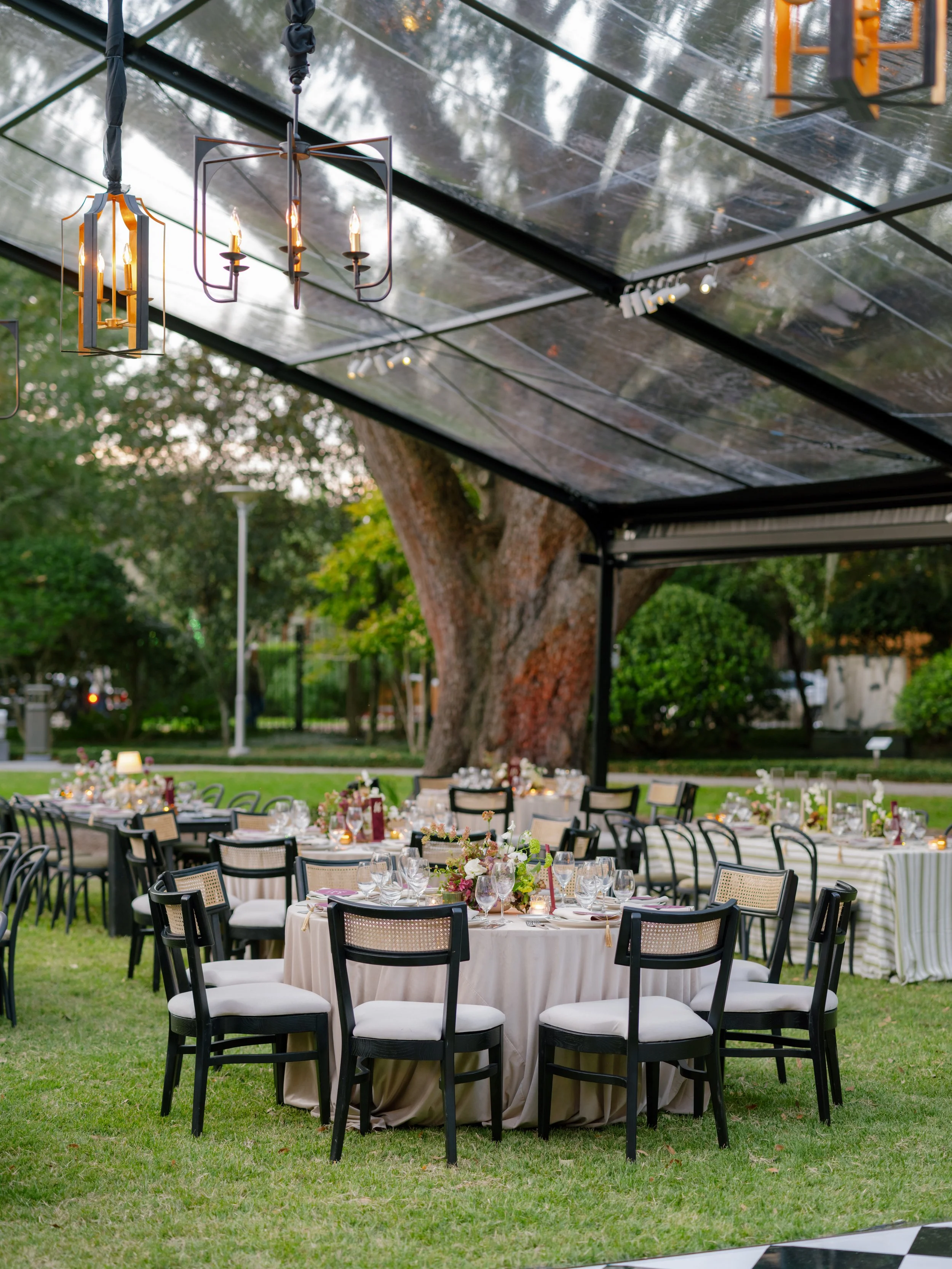 Elegant outdoor event setup with round tables covered in white tablecloths, floral centerpieces, and black chairs under transparent tent with chandeliers overhead, set in a lush green garden with trees.