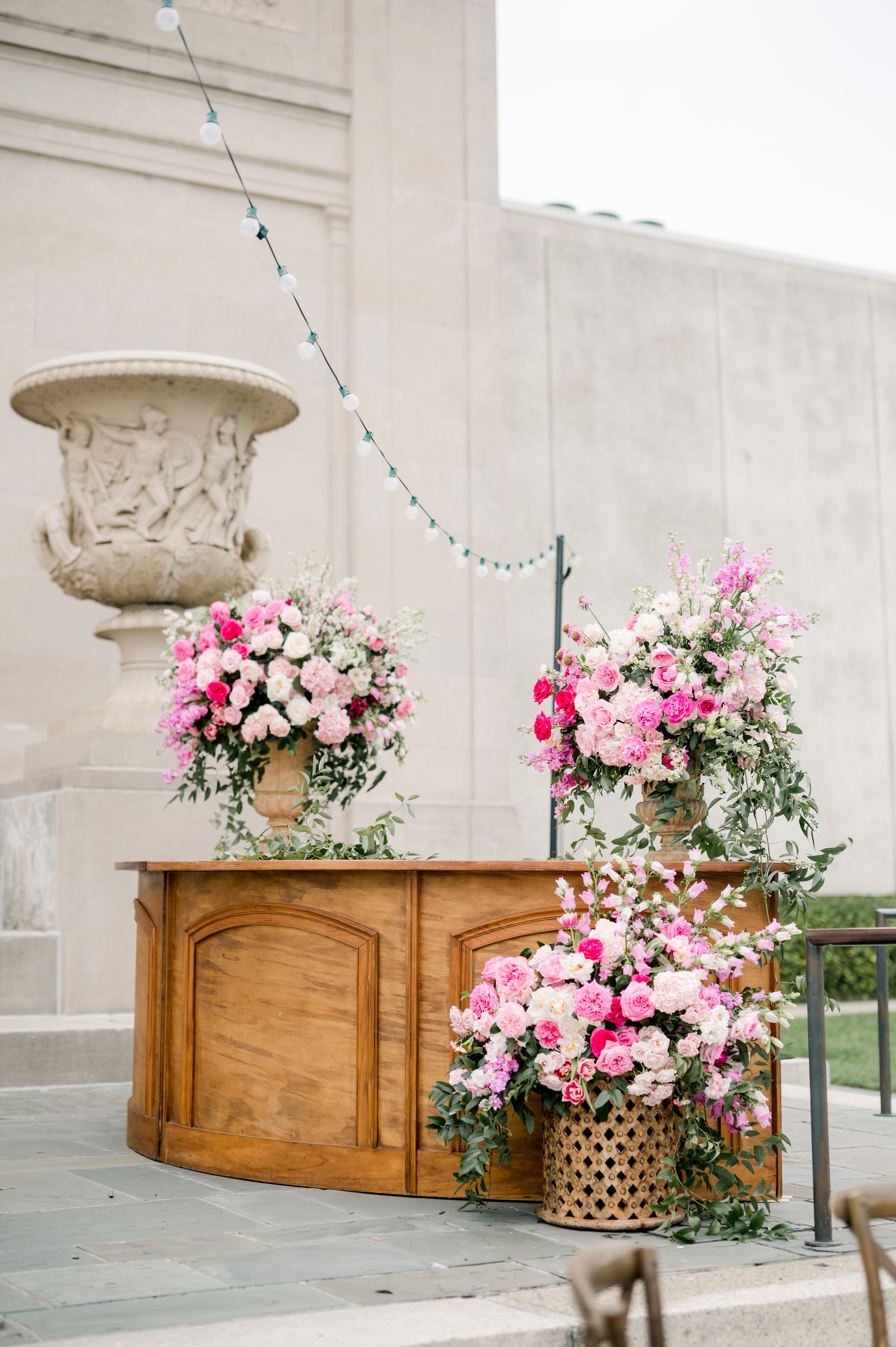 A wooden table with large pink and white flower arrangements in vases, on a tiled outdoor surface, with a decorative stone urn holding a similar floral arrangement and a wall with string lights overhead.