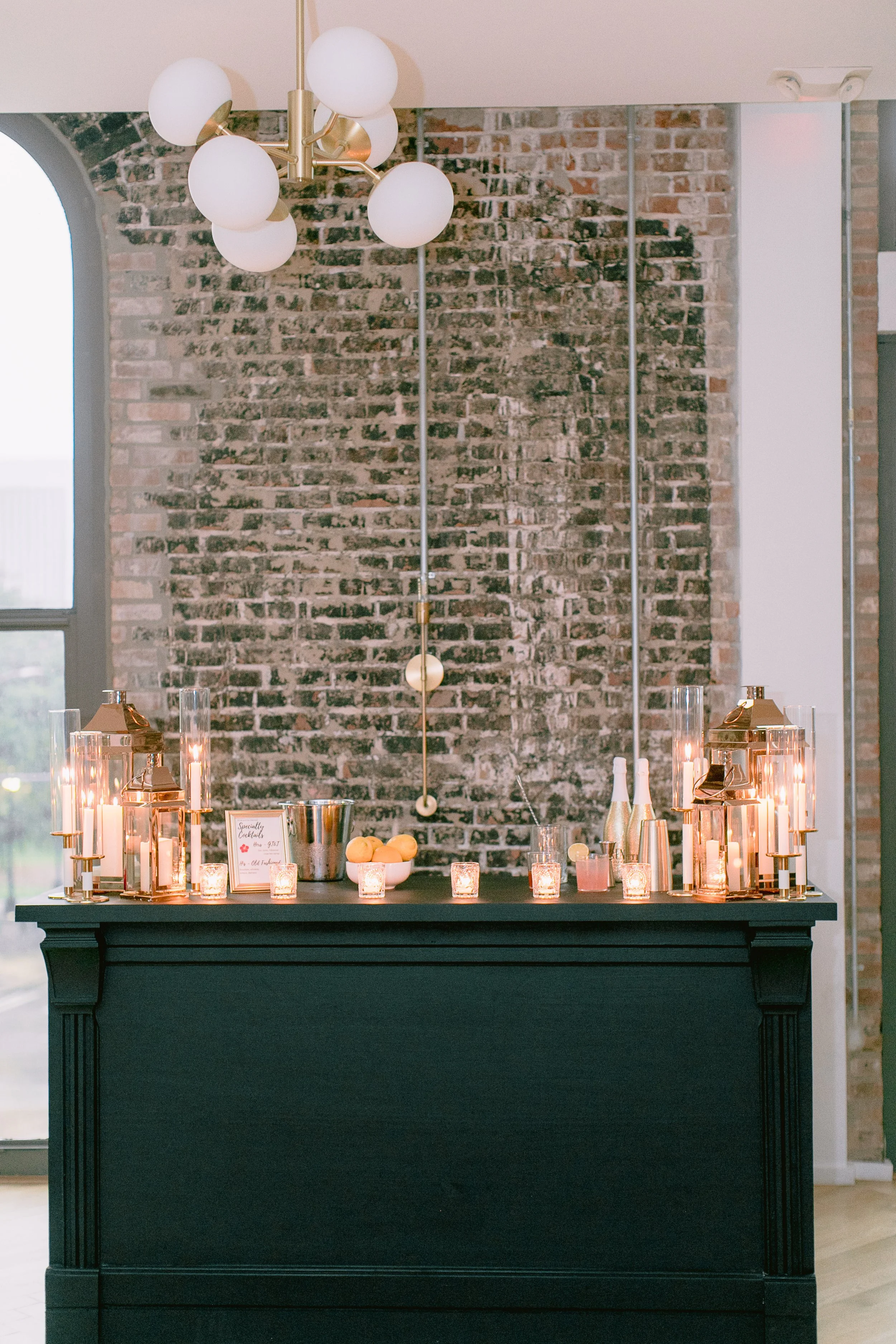 A black table decorated with lit candles, glassware, a bowl of lemons, and a framed sign, set against a brick wall with modern white and gold decor and a chandelier hanging above.