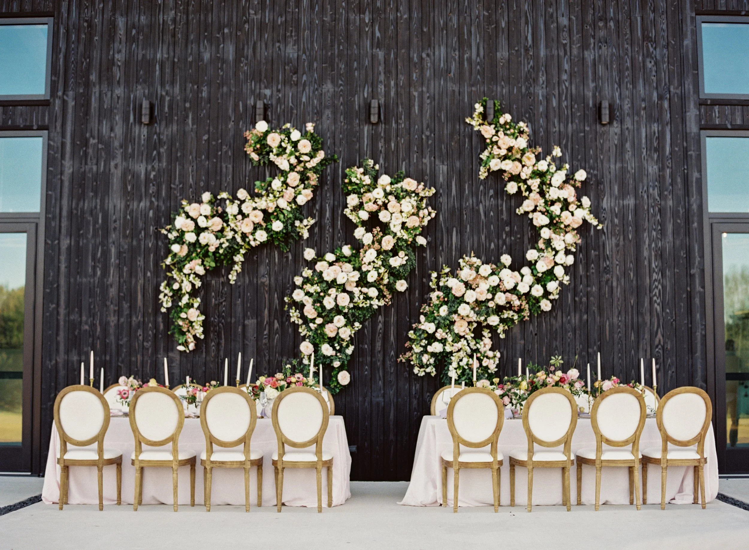 A decorated indoor dining table setup with white tablecloths, surrounded by chairs with white cushions and light wooden frames, featuring floral arrangements. The backdrop is a dark wooden wall with three pink and white floral arrangements attached, 