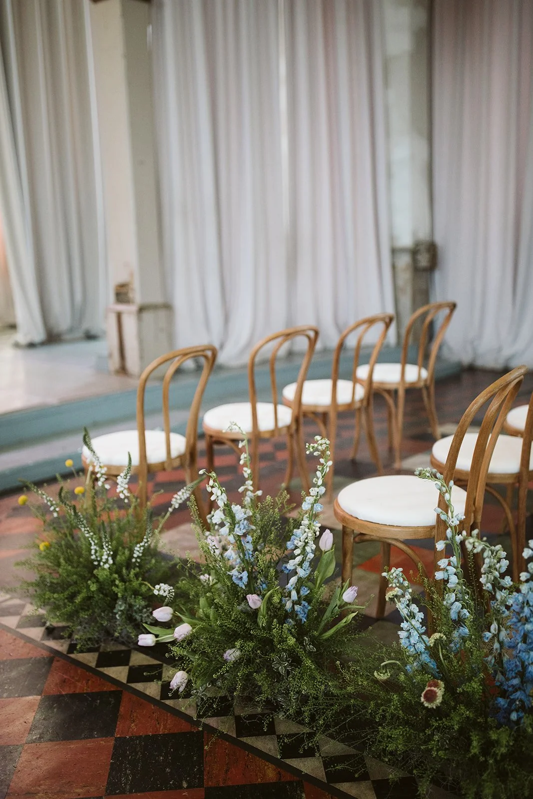Rows of wooden chairs with white cushions arranged on a floral carpet, with white flowers and greenery in front, set against a backdrop of white curtains