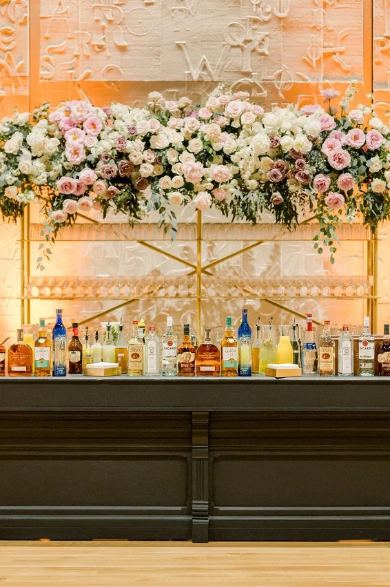 A bar with a large floral arrangement featuring pink and white roses and greenery, set against a textured background with abstract letters.