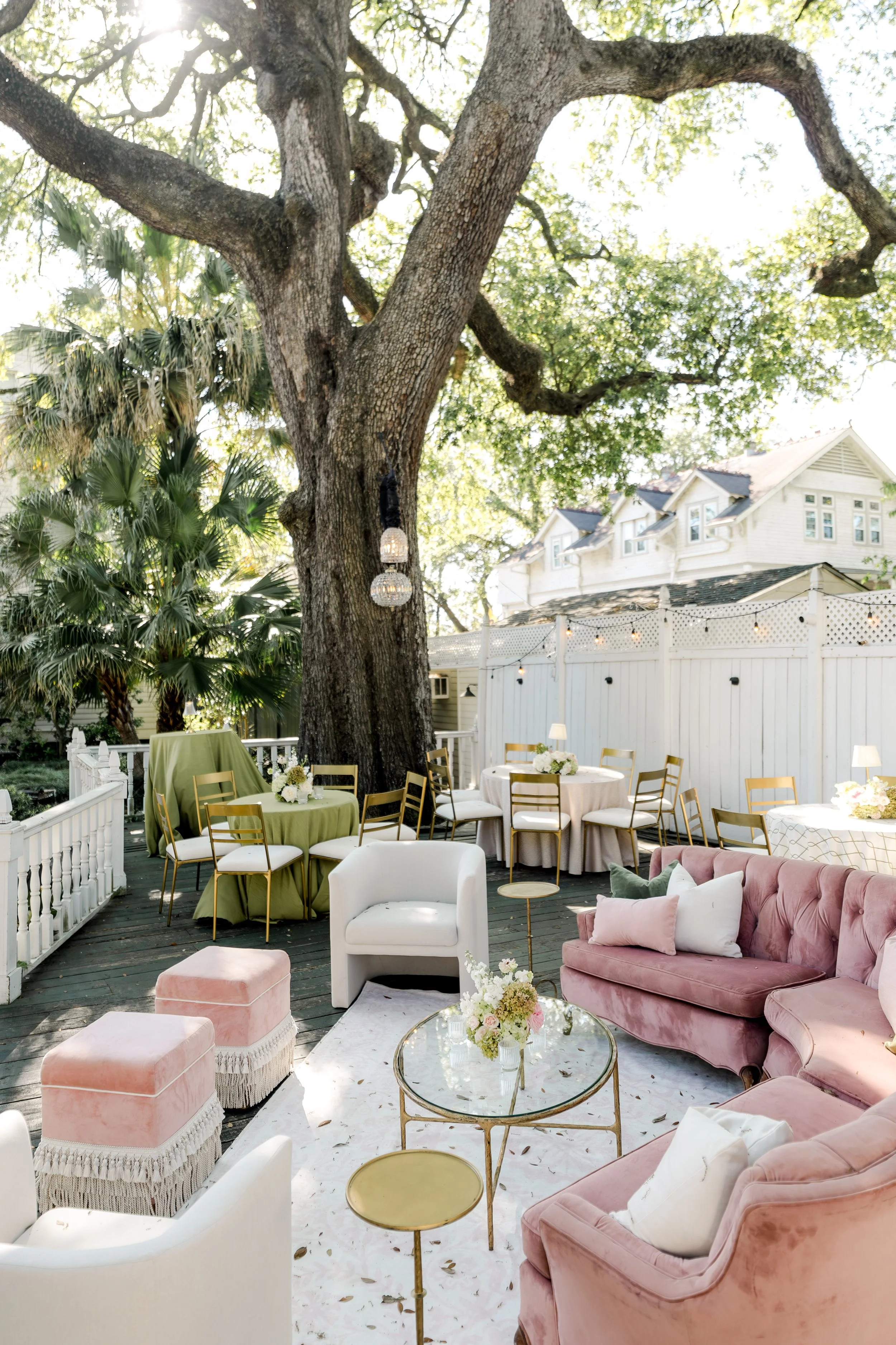 An outdoor patio area decorated for a wedding reception with a pink sectional sofa, coral fringe ottomans, cream lounge chairs and a gold coffee table at Elms Mansion in New Orleans.
