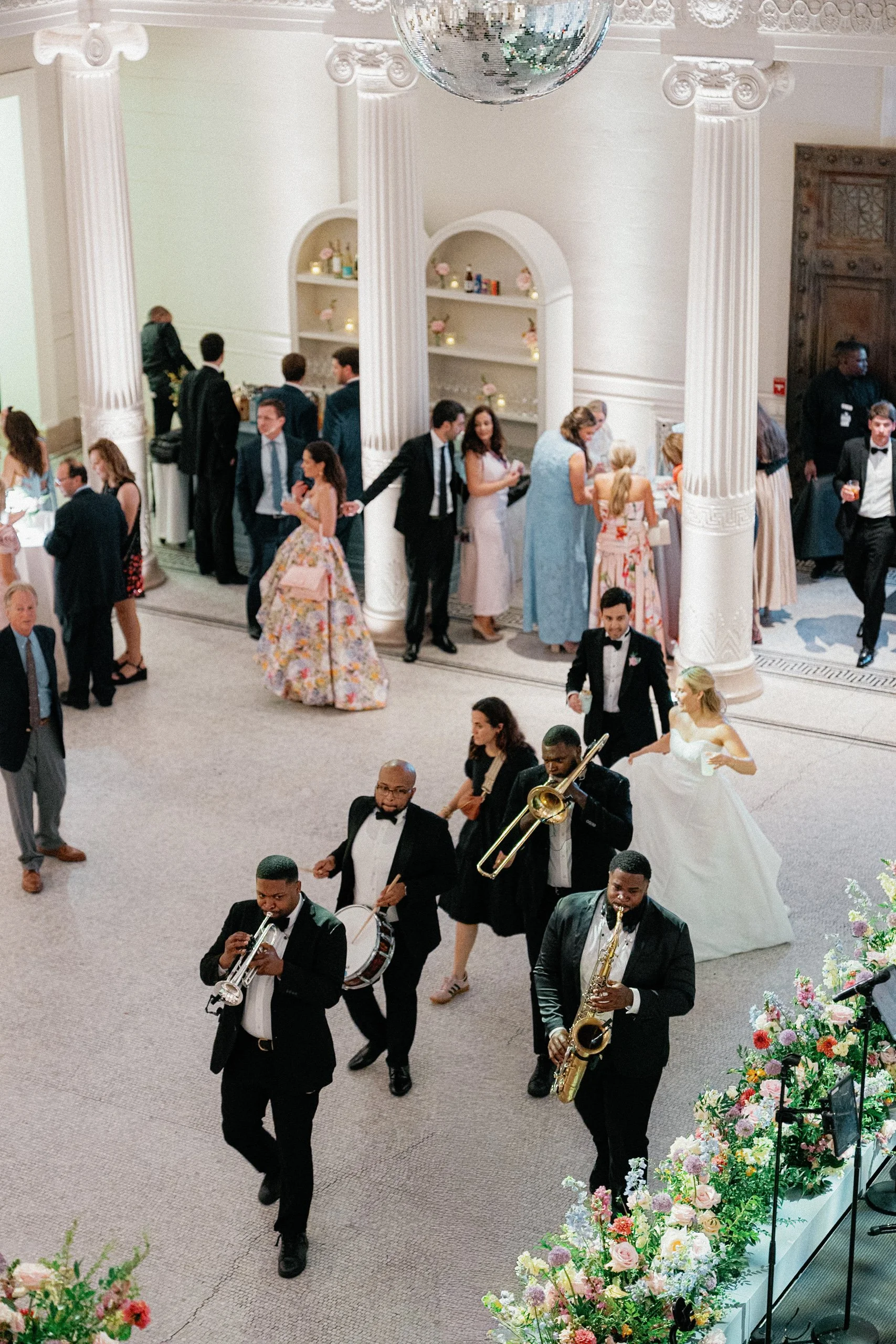 A wedding reception with a marching band playing music, a bride in a white gown, and guests mingling in an elegant decorated hall with white columns, floral arrangements, and a disco ball hanging from the ceiling.