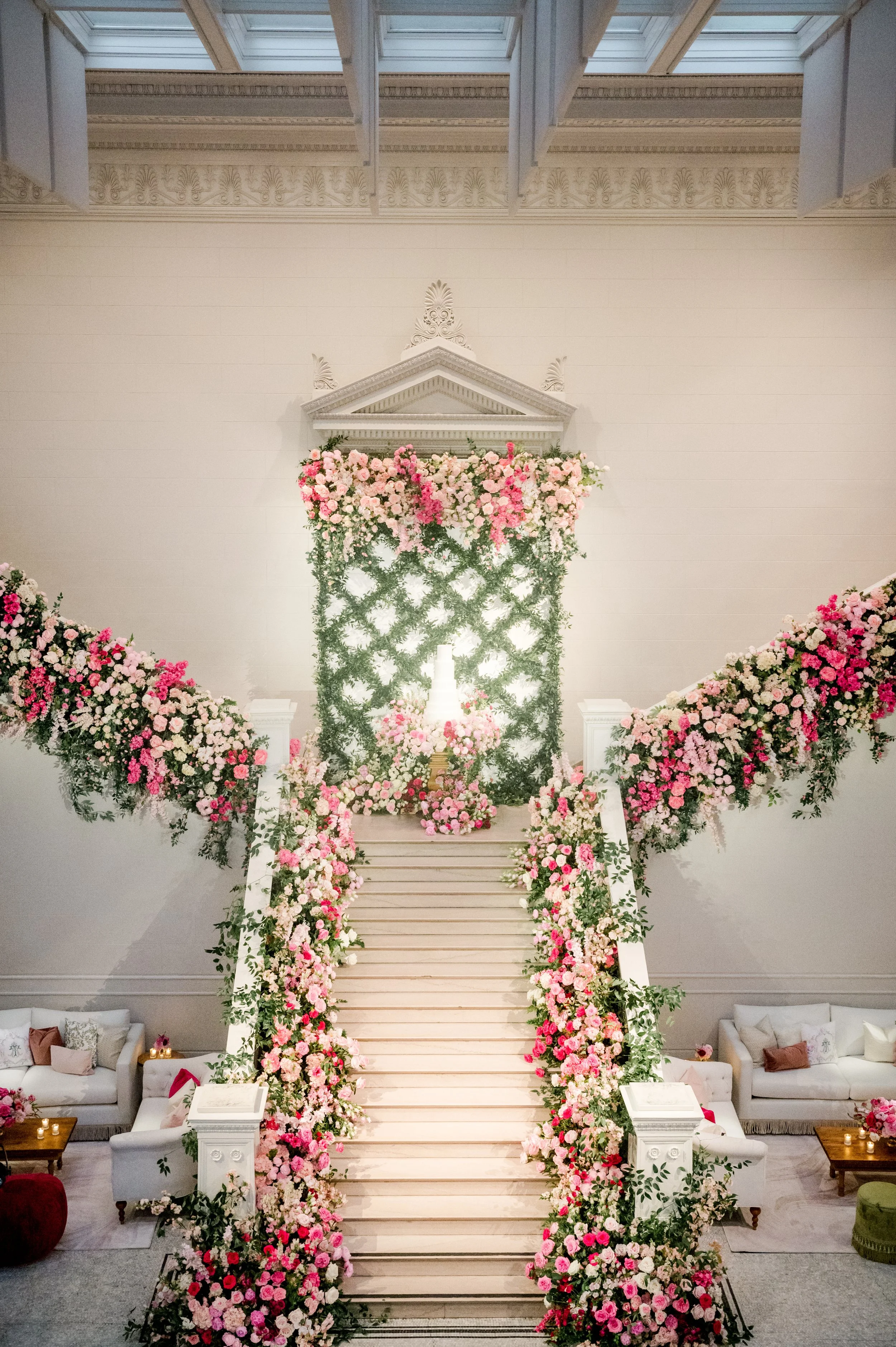 Indoor wedding setup with a staircase decorated with pink and white flowers, leading to a white architectural backdrop with greenery and floral arrangements.