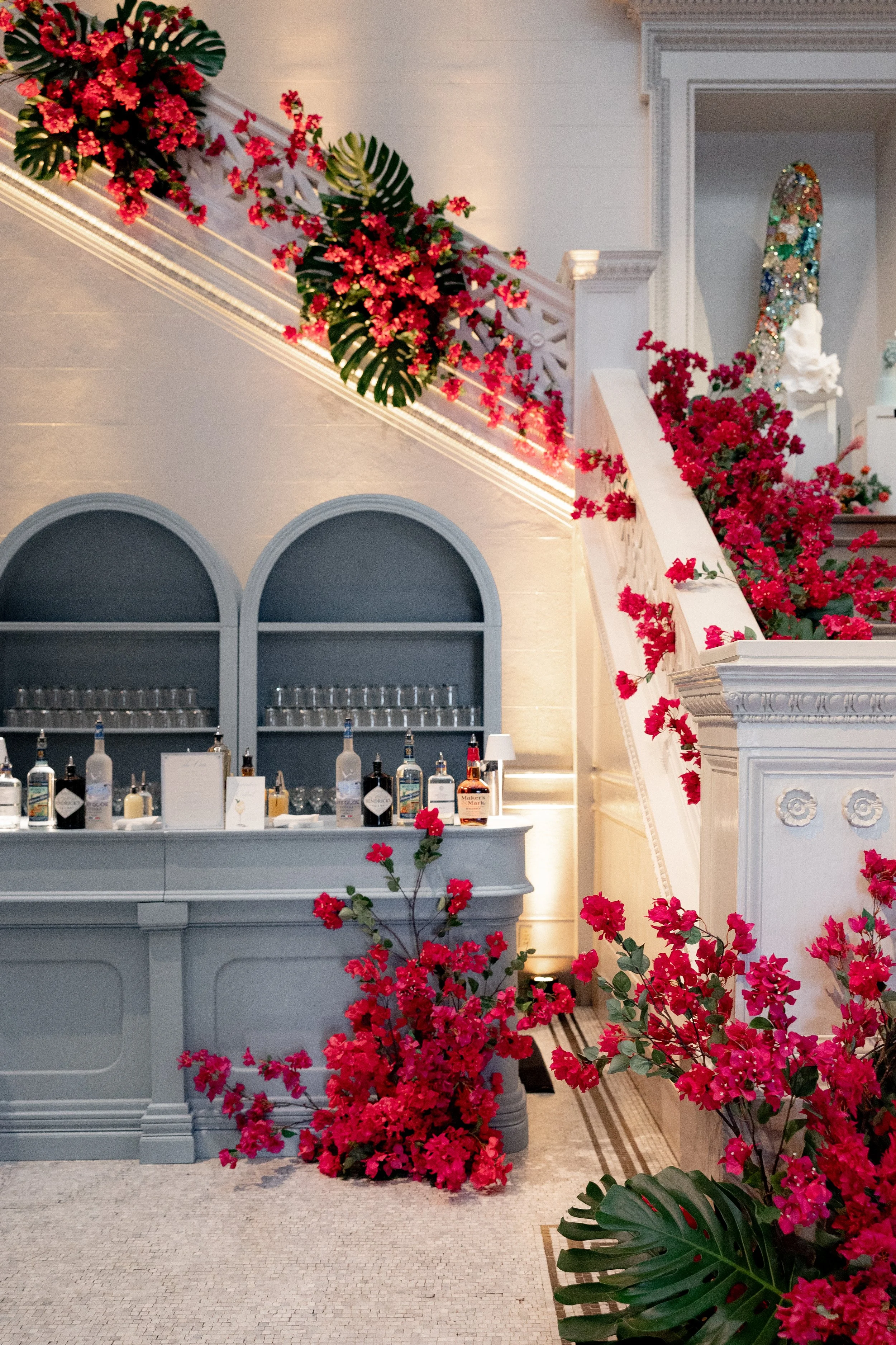 Indoor scene with pink bougainvillea flowers decorating the staircase and a bar area with bottles, glasses, and framed signs, in an elegant, light-colored interior with decorative molding.