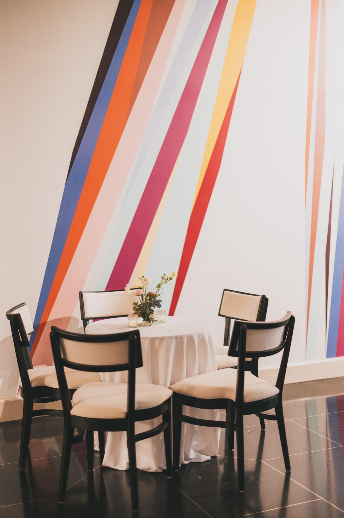 A round table with a white tablecloth, a flower arrangement, and four chairs in a room with a colorful striped wall mural.