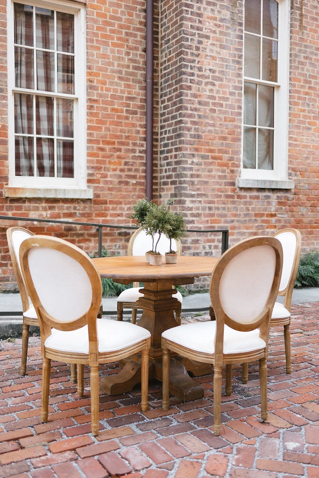 Outdoor patio with a round wooden table and four cream-colored chairs, a potted plant on the table, brick wall background, and large windows.