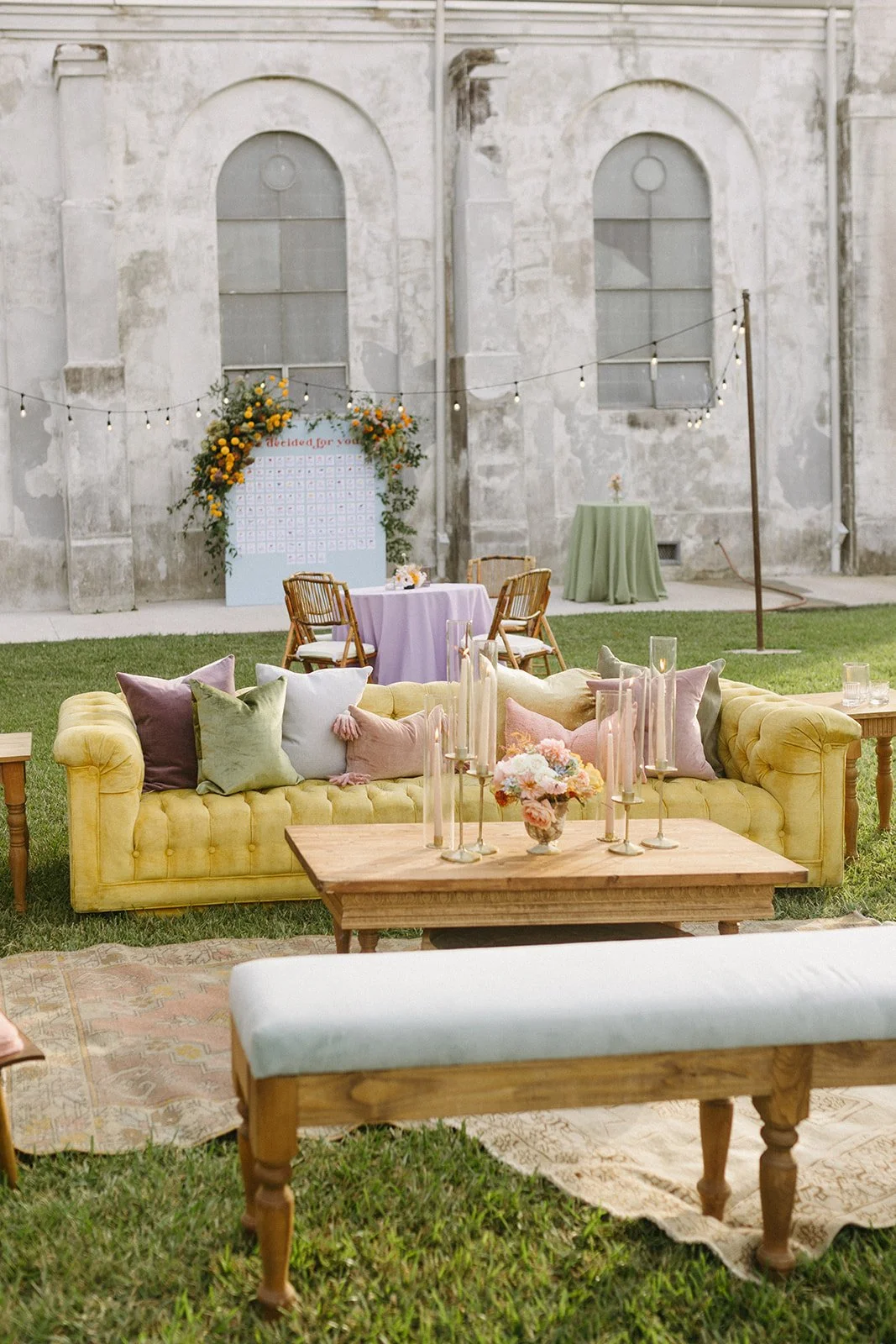 Outdoor wedding or event setup with a yellow tufted sofa, pastel-colored pillows, a wooden coffee table with candles and flowers, a purple-draped table with chairs in the background, string lights, and a decorative board with yellow flowers against a