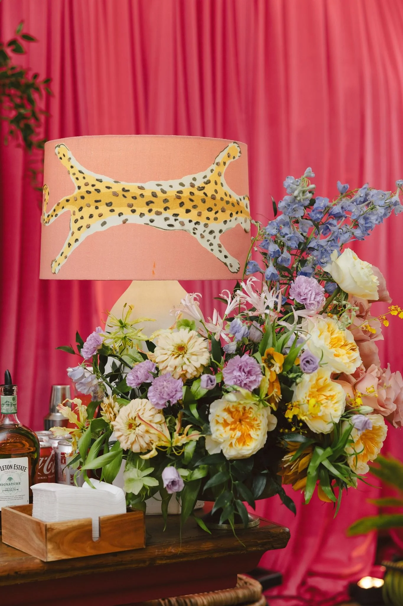 A pink table lamp with a giraffe pattern sits amid a large colorful floral arrangement on a wooden table. The background features pink curtains.