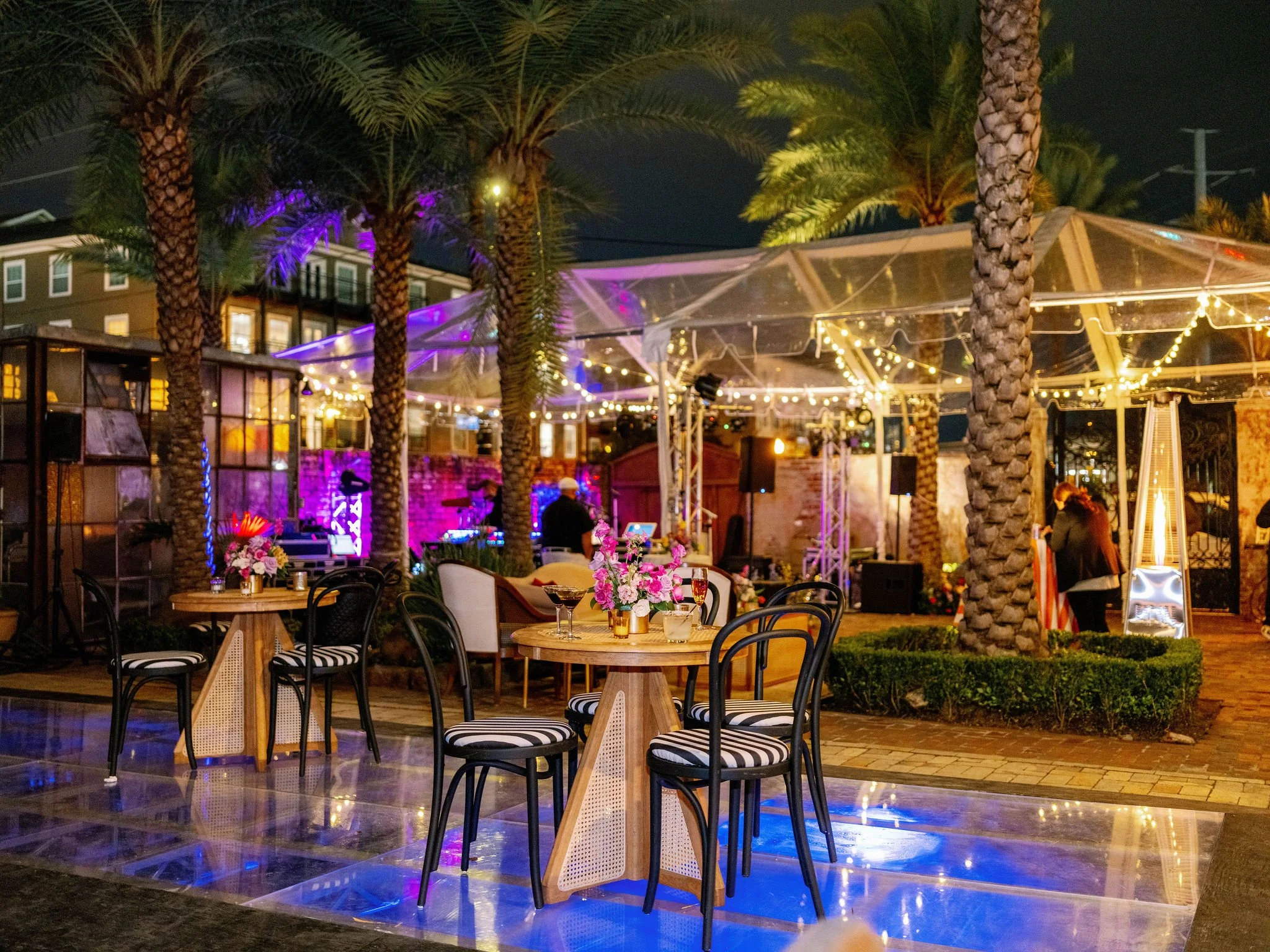 Outdoor restaurant patio at night with tables, chairs, floral centerpieces, string lights, palm trees, and a stage with purple lighting.
