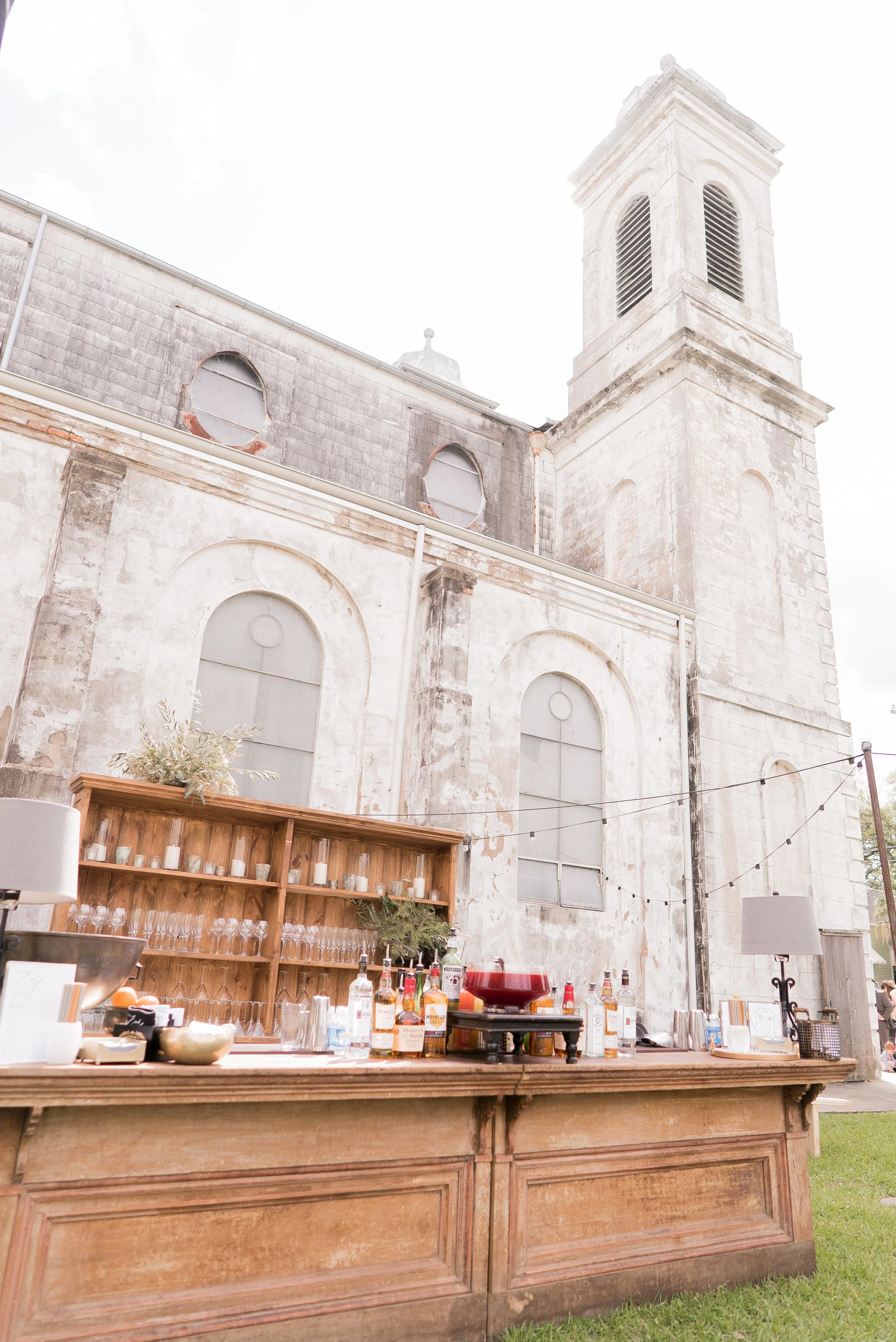 An outdoor bar setup in front of an old church with a tall bell tower. The bar has a wooden counter with various bottles of alcohol, glasses, and barware. The church has a weathered exterior with arched windows and a clock face. Some string lights ar