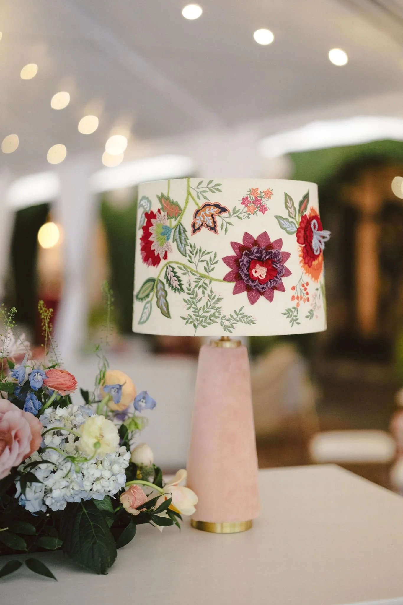 A floral lamp with a cylindrical shade featuring colorful embroidered flowers, placed on a table with a pale pink base and gold accents, surrounded by a bouquet of various fresh flowers.