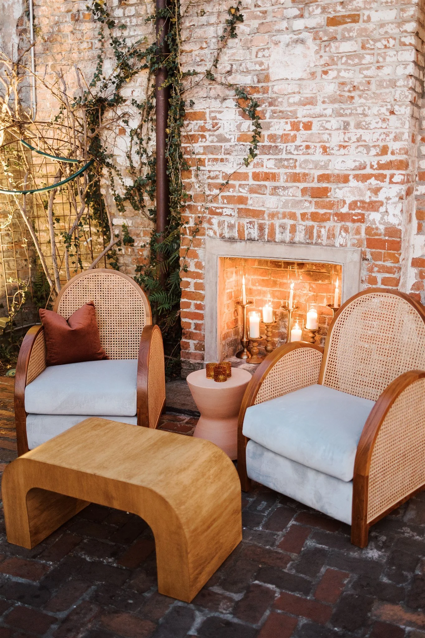 Outdoor seating area with two white cushioned chairs with rattan frames and brown pillows, a wooden coffee table, and a small side table with candles. The background features a brick fireplace with lit candles inside, a brick wall, some green ivy and