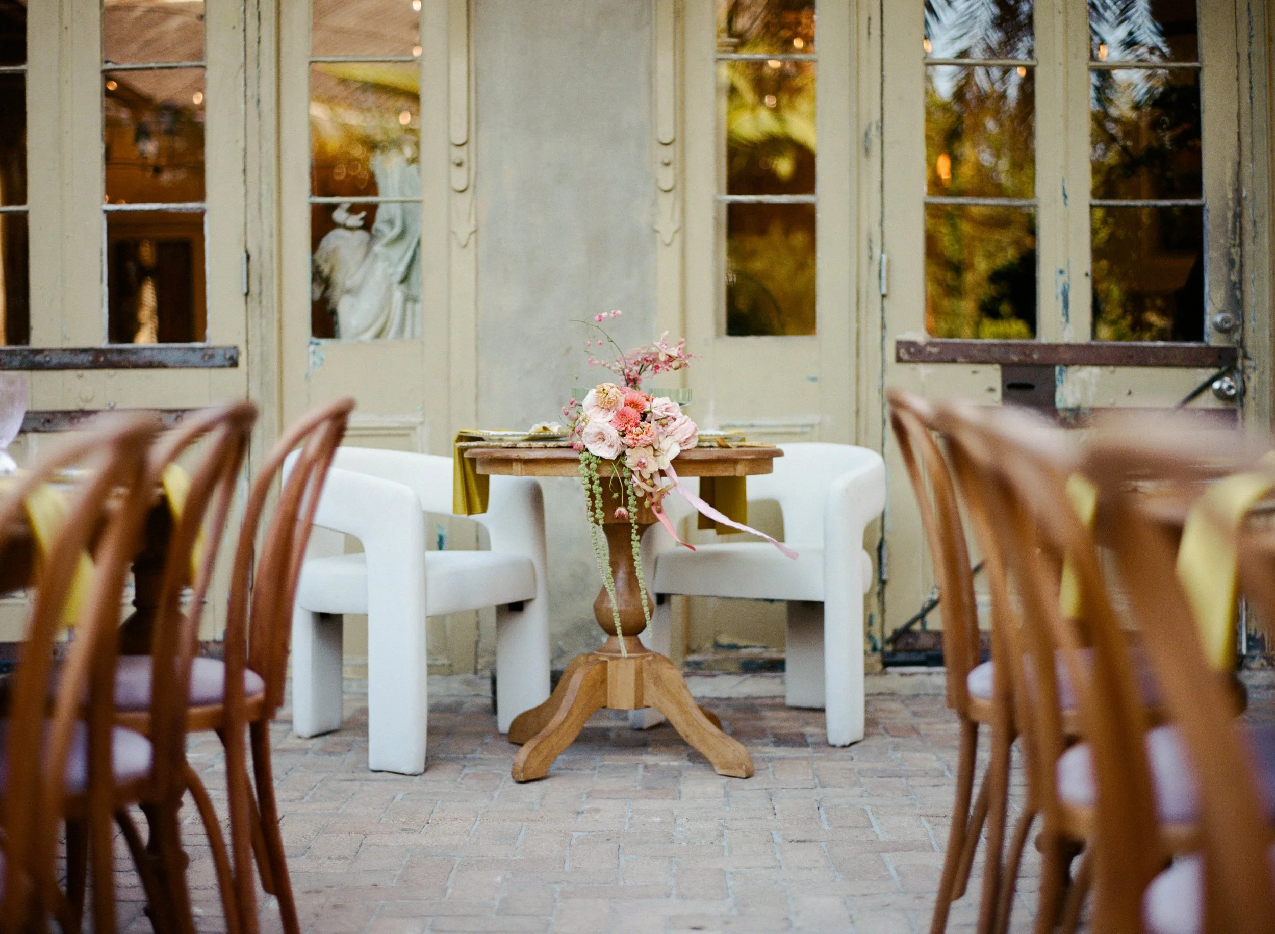 A dining area with a wooden table decorated with a pink floral centerpiece, surrounded by white chairs. Behind the table are large glass doors with multiple panes, revealing a garden with trees.