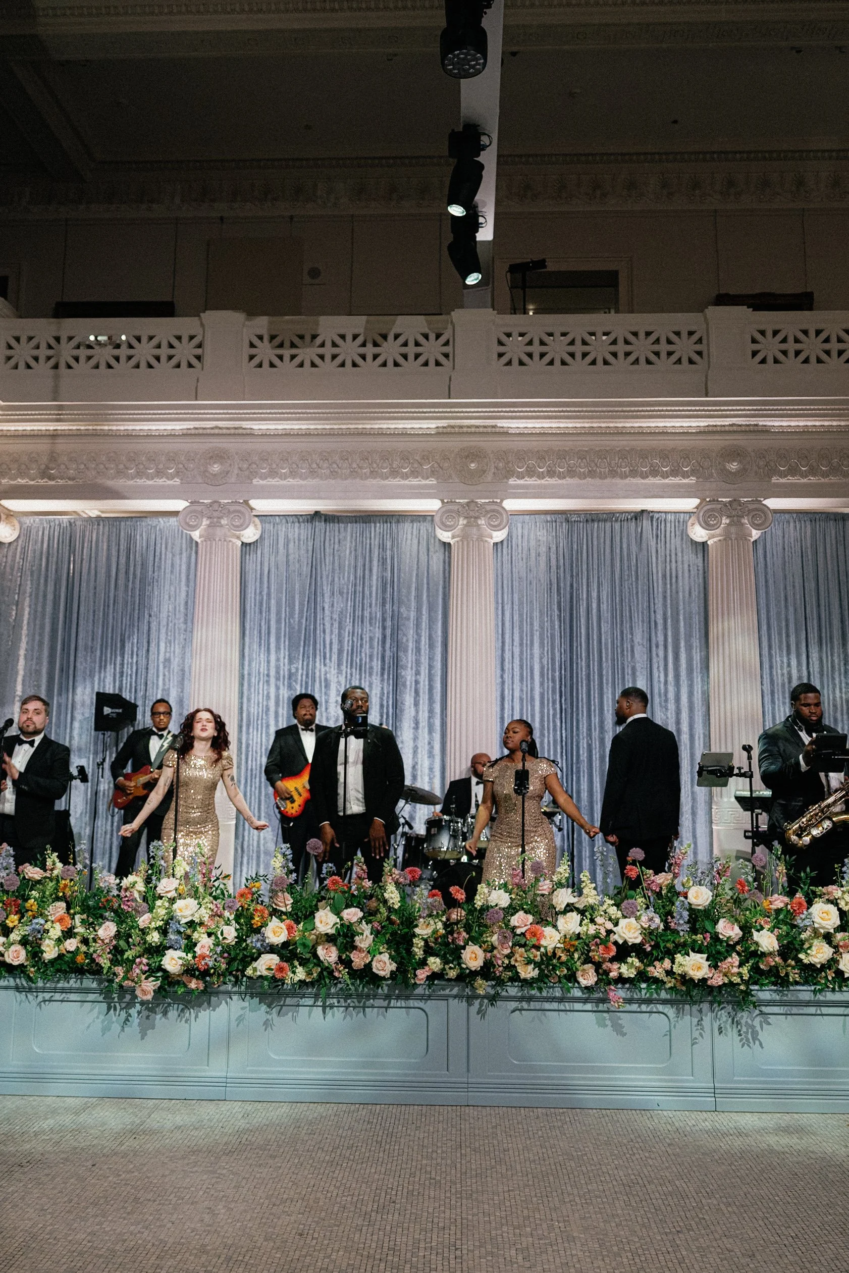 A group of musicians and singers performing on stage with a flower arrangement in front, in a grand, decorated hall.