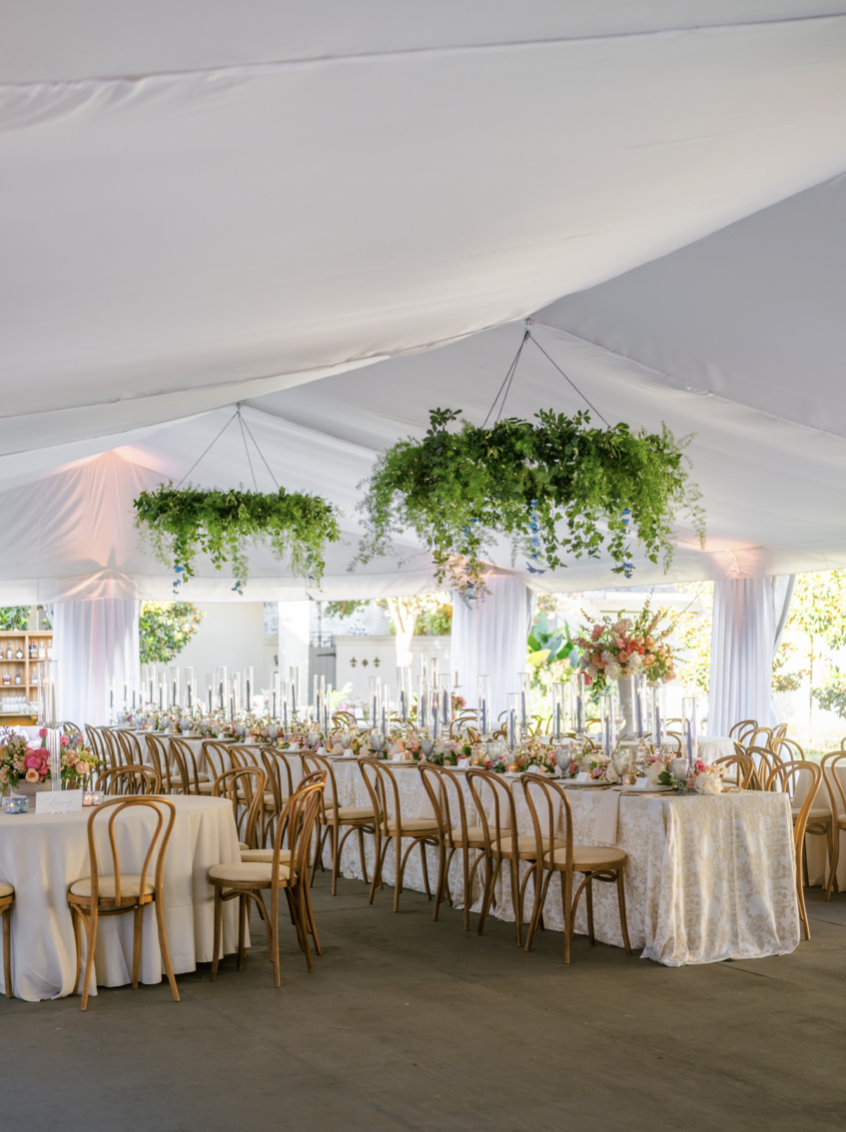 Elegant wedding reception setup with round and rectangular tables, floral centerpieces, and hanging green plants inside a white tent.