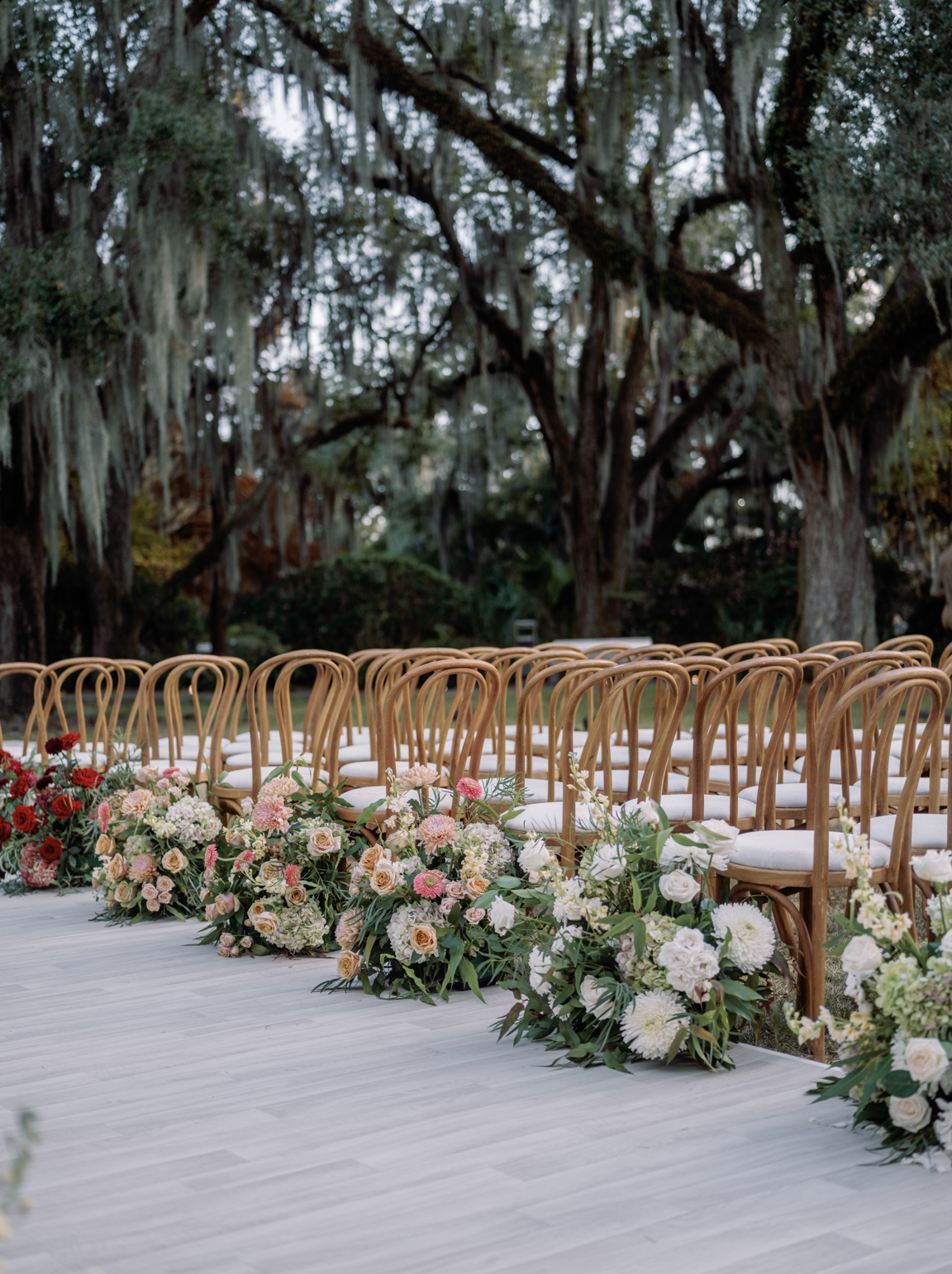Outdoor wedding ceremony setup with wooden chairs and floral arrangements on a white aisle runner in a garden with large moss-covered trees.