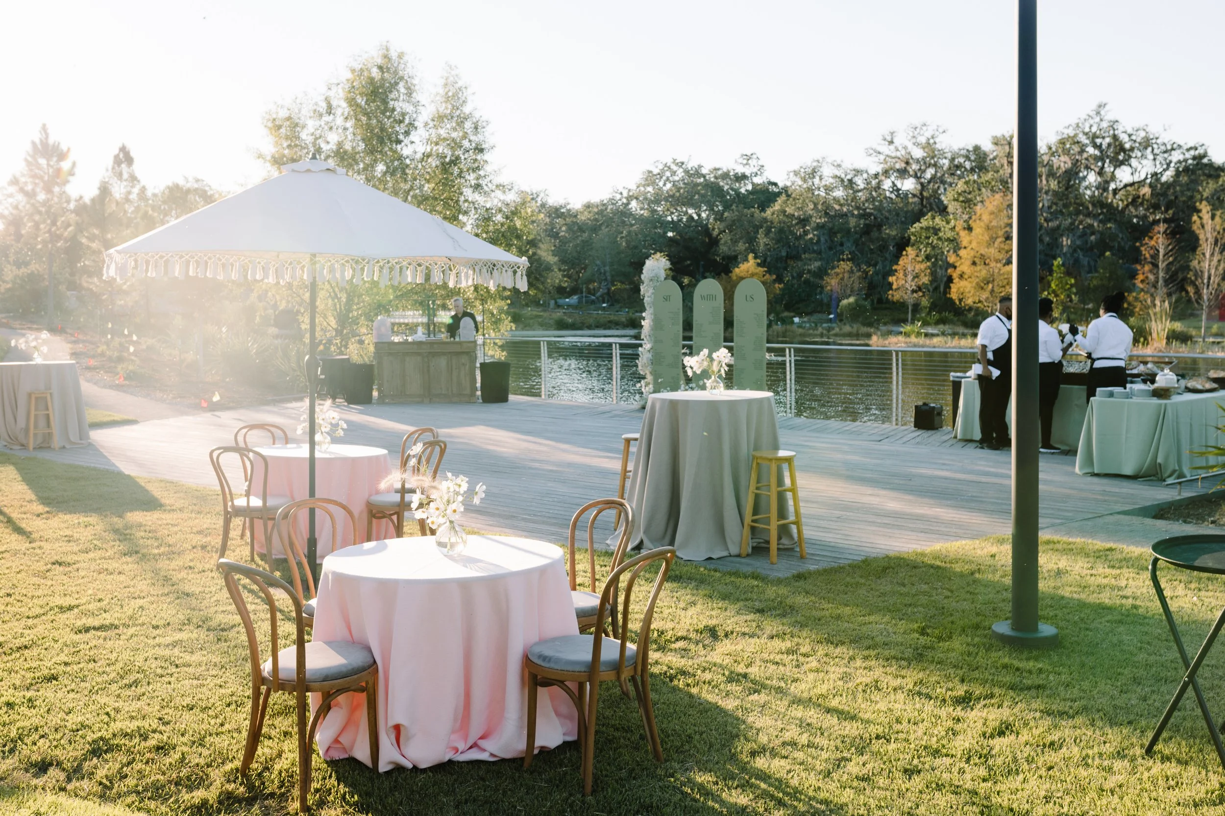 An outdoor event setup near a lake with round tables covered in pink tablecloths, wooden chairs, a white parasol, and a buffet area with staff preparing food. The scene is bathed in warm sunlight and surrounded by trees.