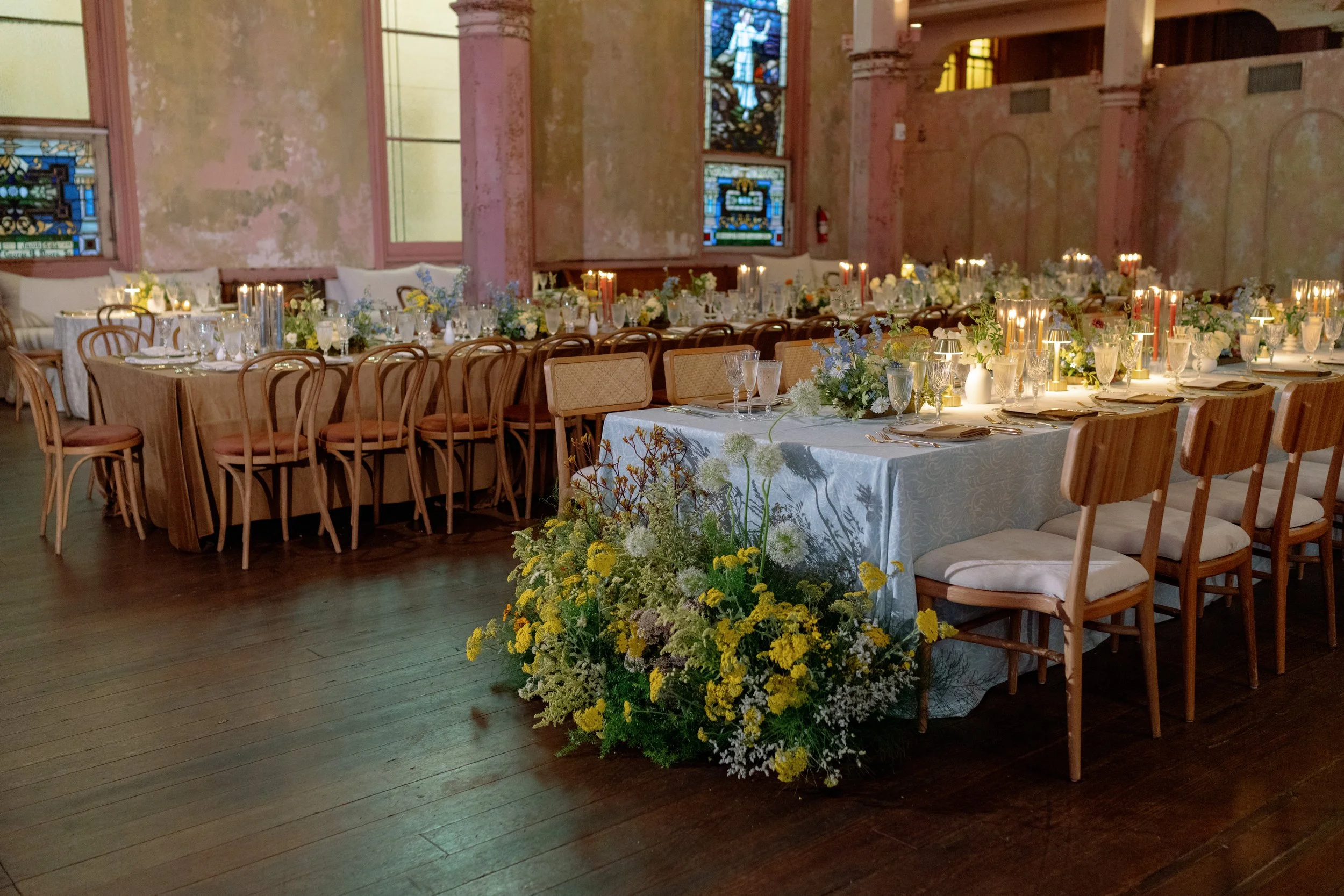 Elegant indoor venue with long dining tables decorated with white tablecloths, floral centerpieces, numerous glasses, and candles. The walls feature stained glass windows and are weathered, pastel-colored with peeling paint. The floor is wooden, and 