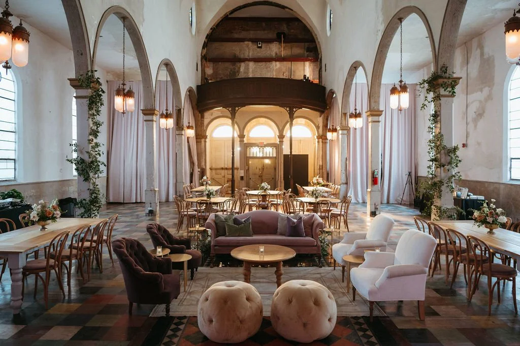 Elegant, spacious event hall with high vaulted ceilings, arched windows, and colonial architecture. Decor includes modern and vintage furniture, floral centerpieces, and soft lighting for a gathering or celebration.