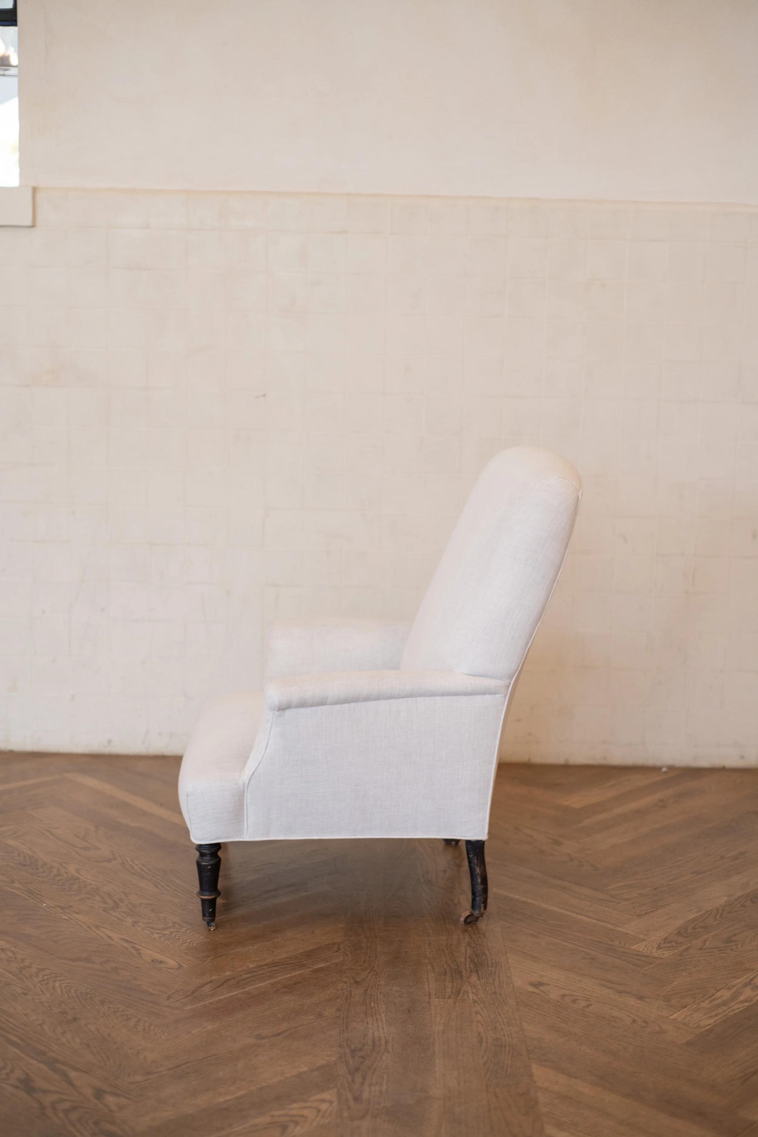 A light beige upholstered armchair with dark wooden legs sitting on a brown wooden floor against a white painted brick wall.