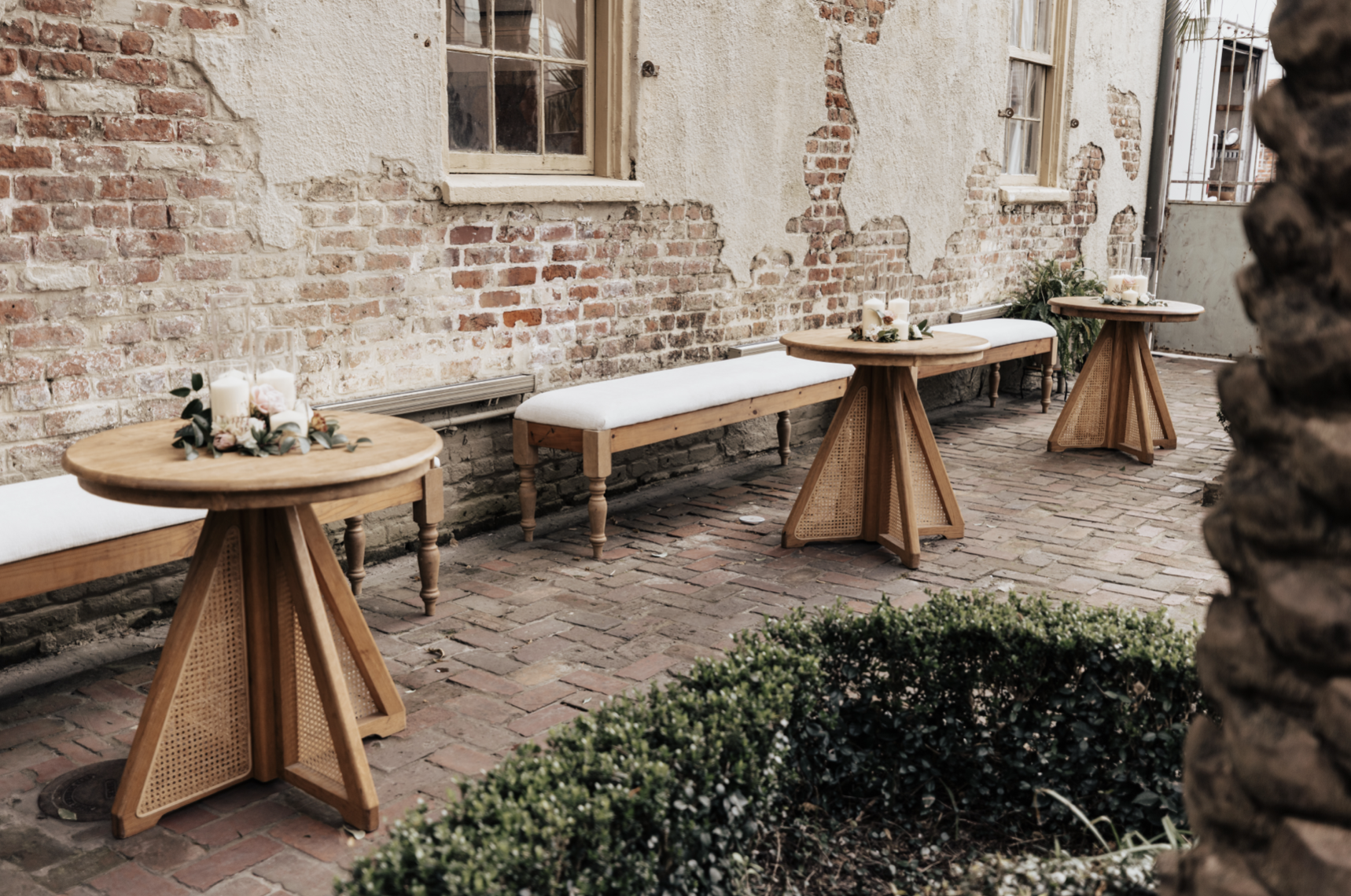Outdoor patio with brick paving, featuring three wooden tables decorated with candles and flowers, and paired with benches against an exposed brick wall.