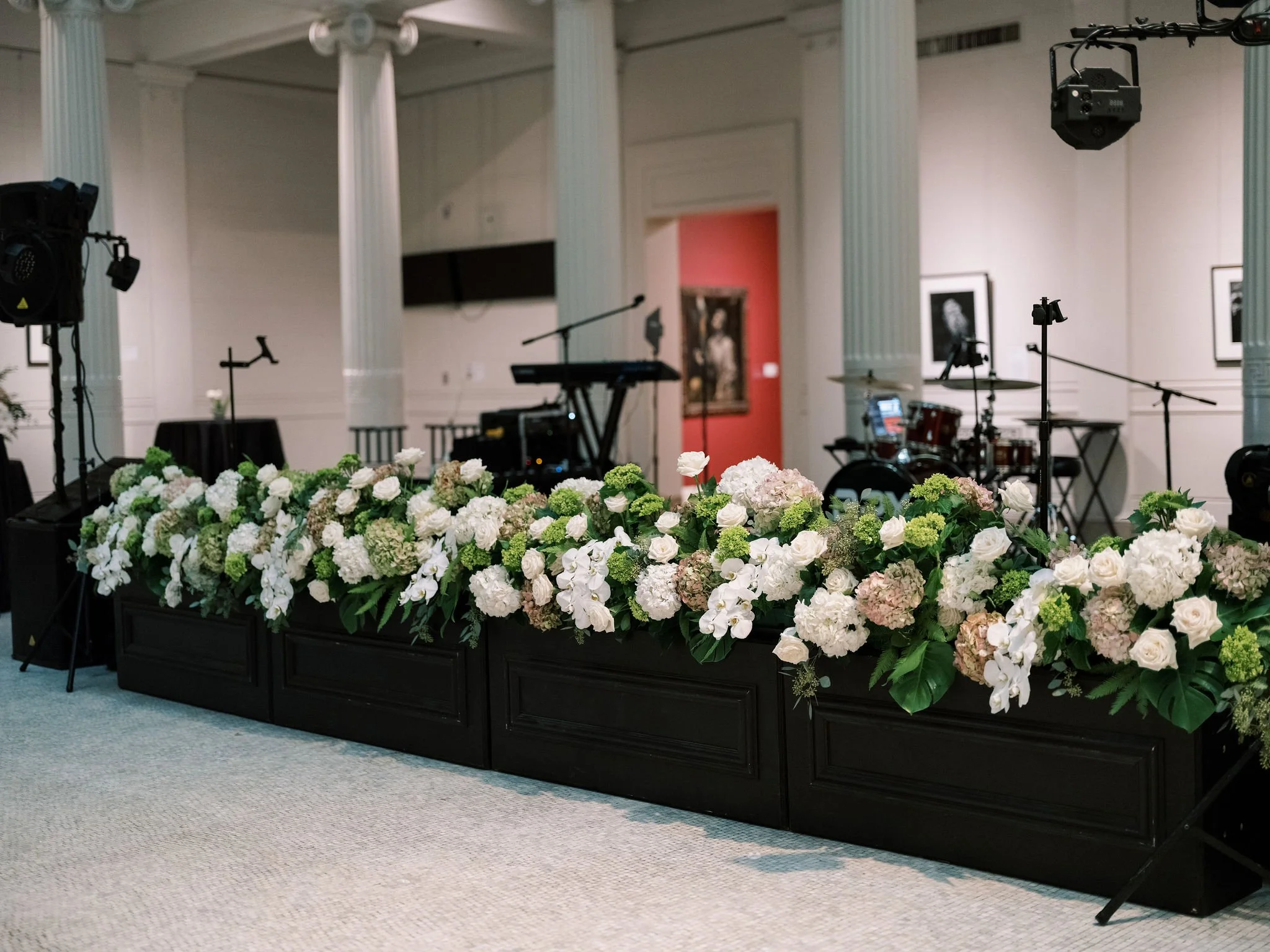 A floral arrangement of white and pale pink flowers decorates a black table in front of a stage with musical instruments in an elegant hall.
