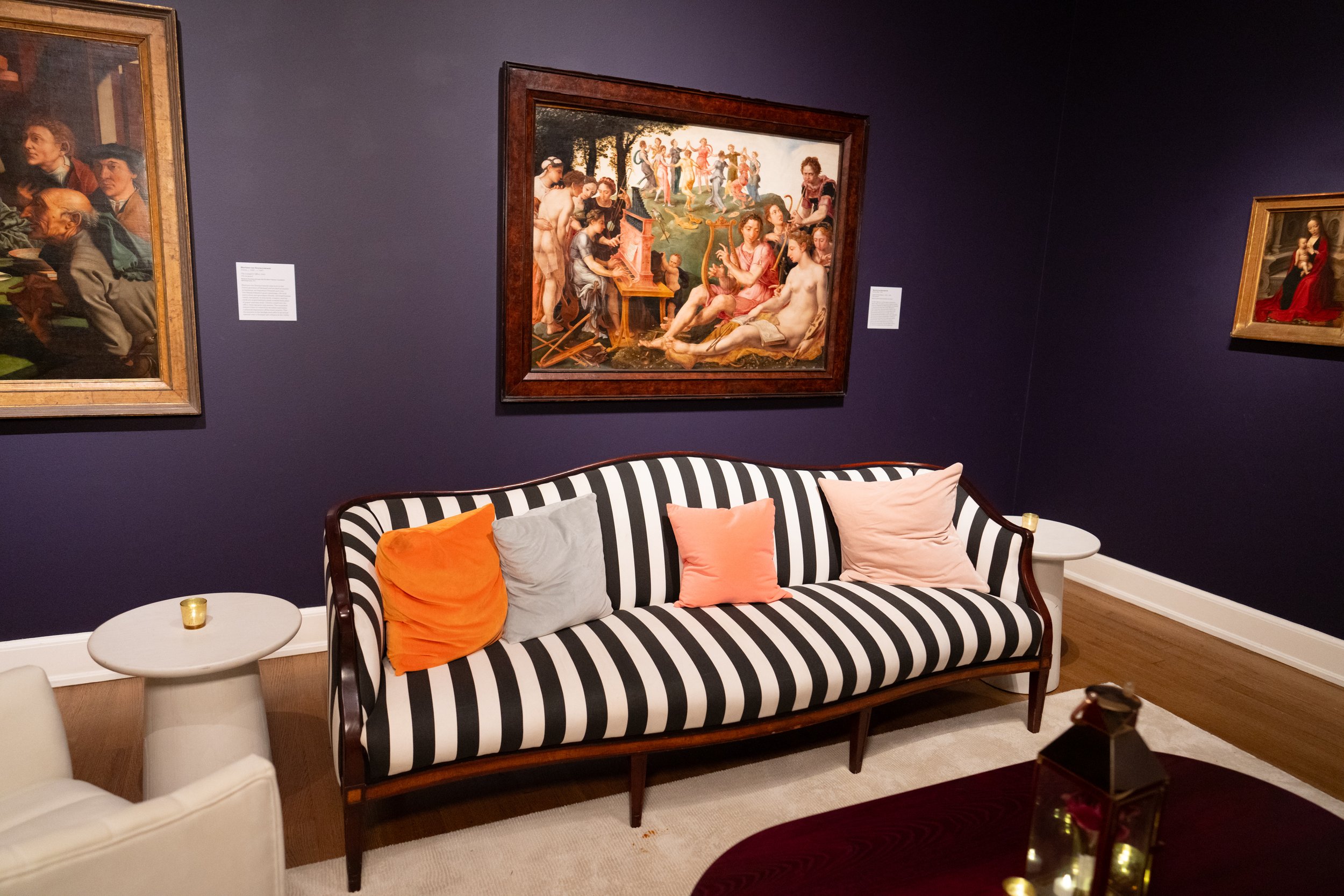 A living room with dark blue walls, a black and white striped sofa with colorful pillows, two small round white tables, a white chair, and framed artworks on the wall including a large painting of people dancing and playing instruments.