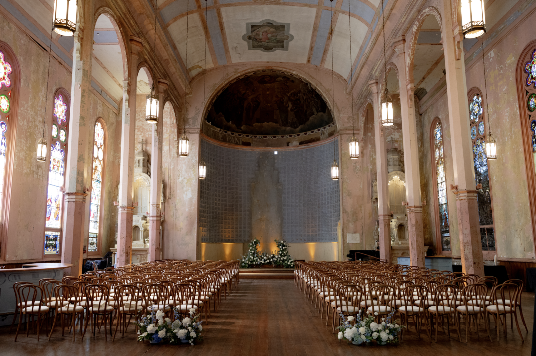 Interior of a church decorated for a wedding with rows of wooden chairs, floral arrangements at the front, stained glass windows on both sides, and a painted ceiling.