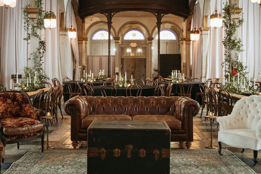 Elegant wedding or event reception hall with vintage-style furniture, round tables with black tablecloths, candle centerpieces, chandeliers, and large windows with curtains, decorated with greenery and candles.