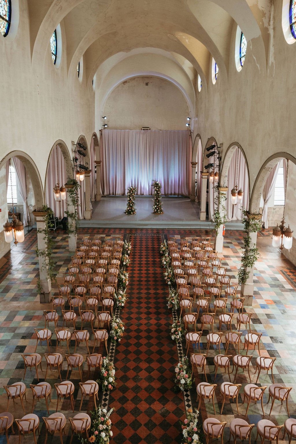 Interior of a wedding or event hall decorated with floral arrangements along the aisle, with rows of chairs facing a stage with a curtain backdrop and floral decor, featuring arched windows and vintage lighting.