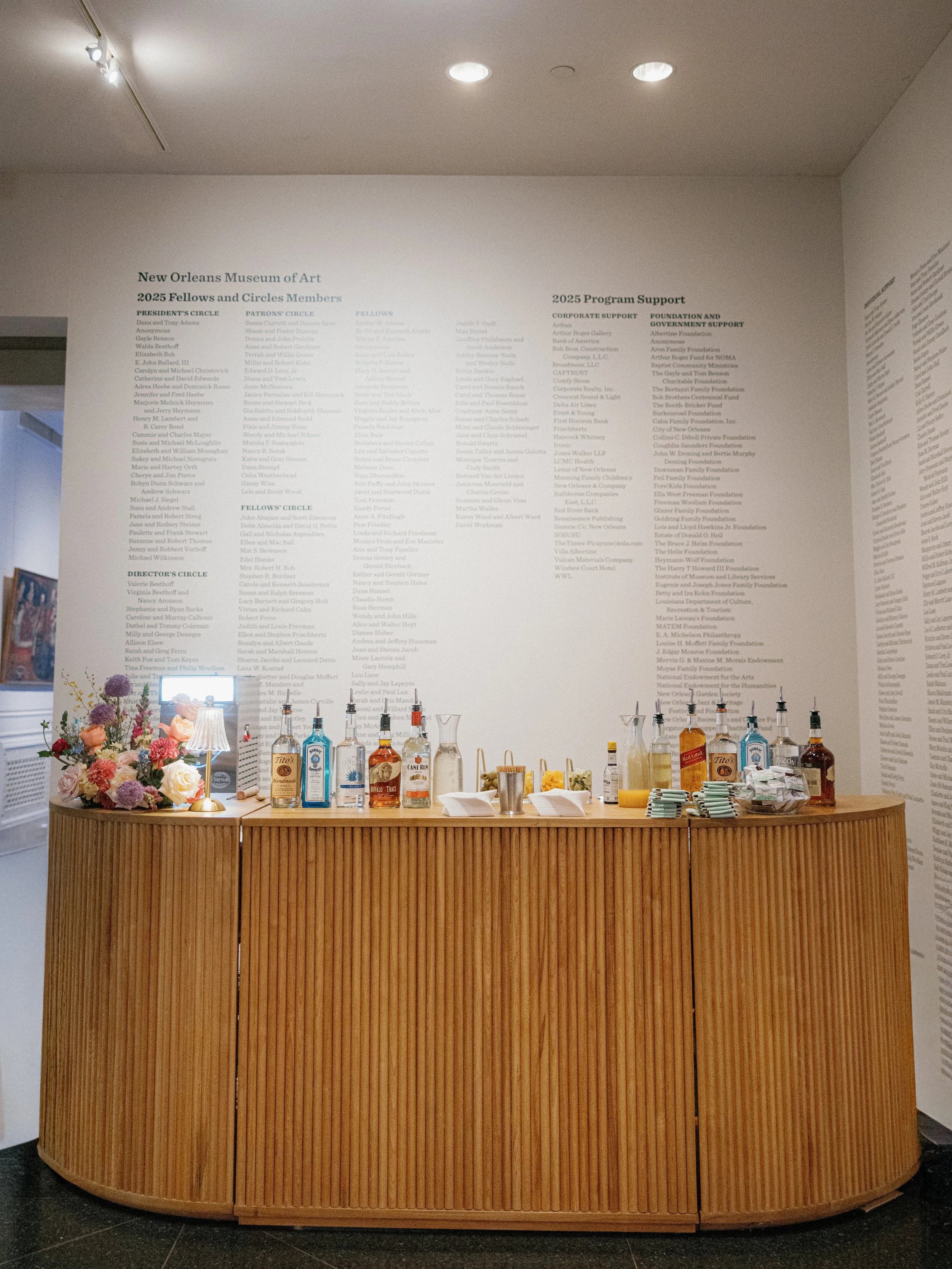 A reception or bar area with a wooden curved counter, several bottles of alcohol, a small stack of napkins, and a floral arrangement. Behind the counter is a white wall with printed lists of donors, sponsors, and supporters for the New Orleans Museum
