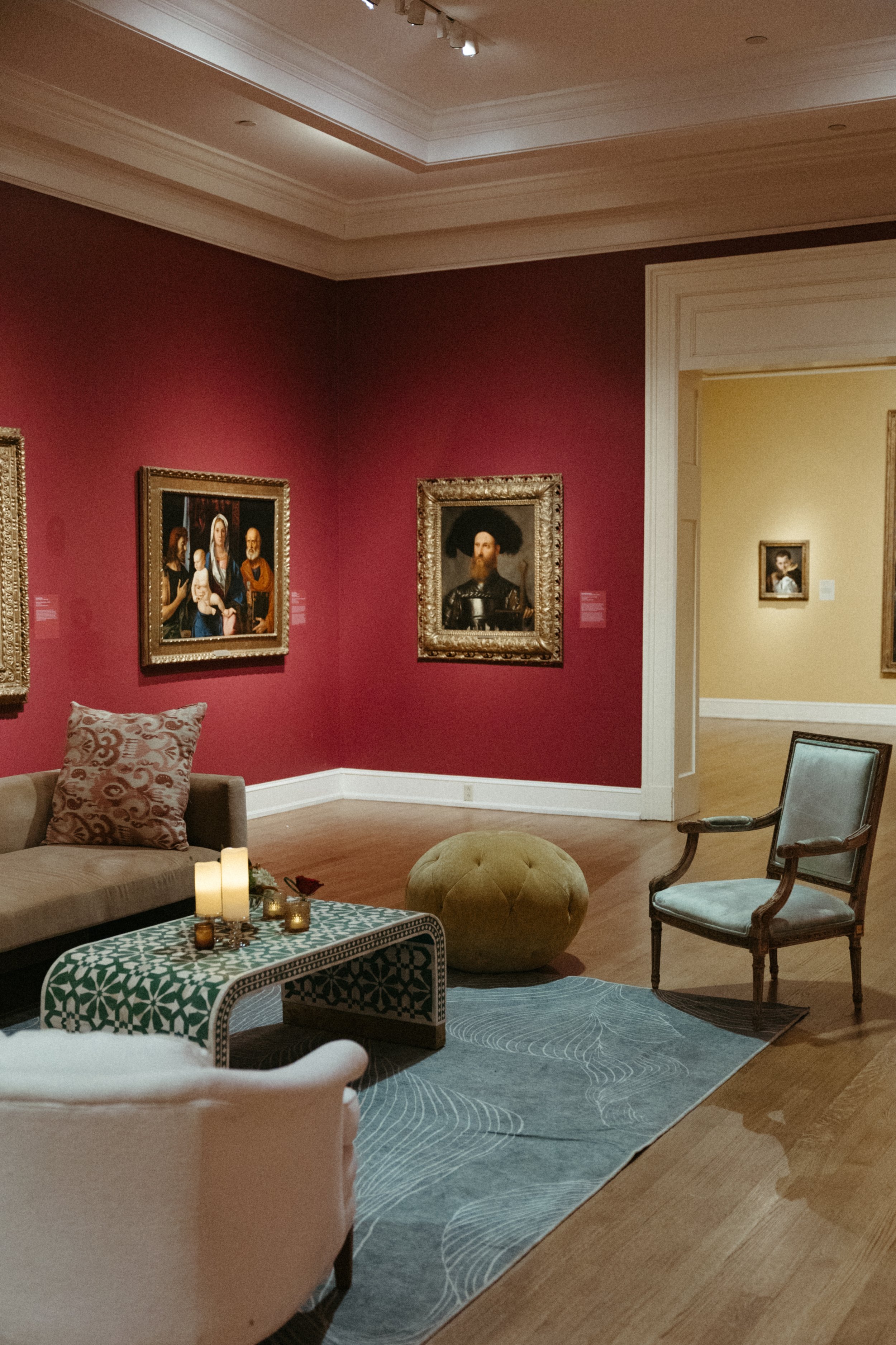 An art gallery room featuring framed classical paintings on pink and beige walls, with various chairs and a small table decorated with candles and a patterned tablecloth.